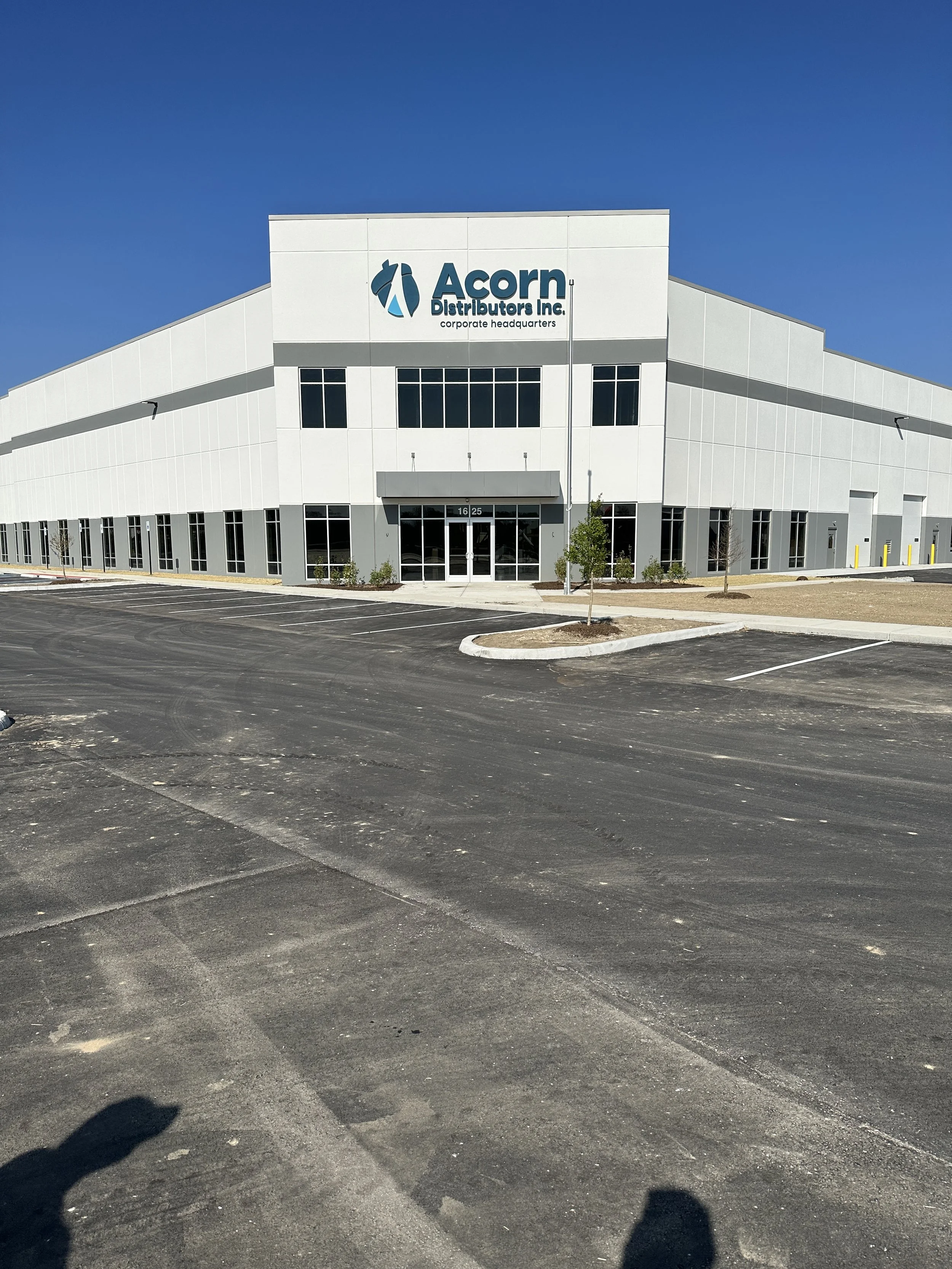 New industrial warehouse building with the sign 'Acorn Distributors Inc. corporate headquarters' on the front, surrounded by a parking lot and small trees under a clear blue sky.