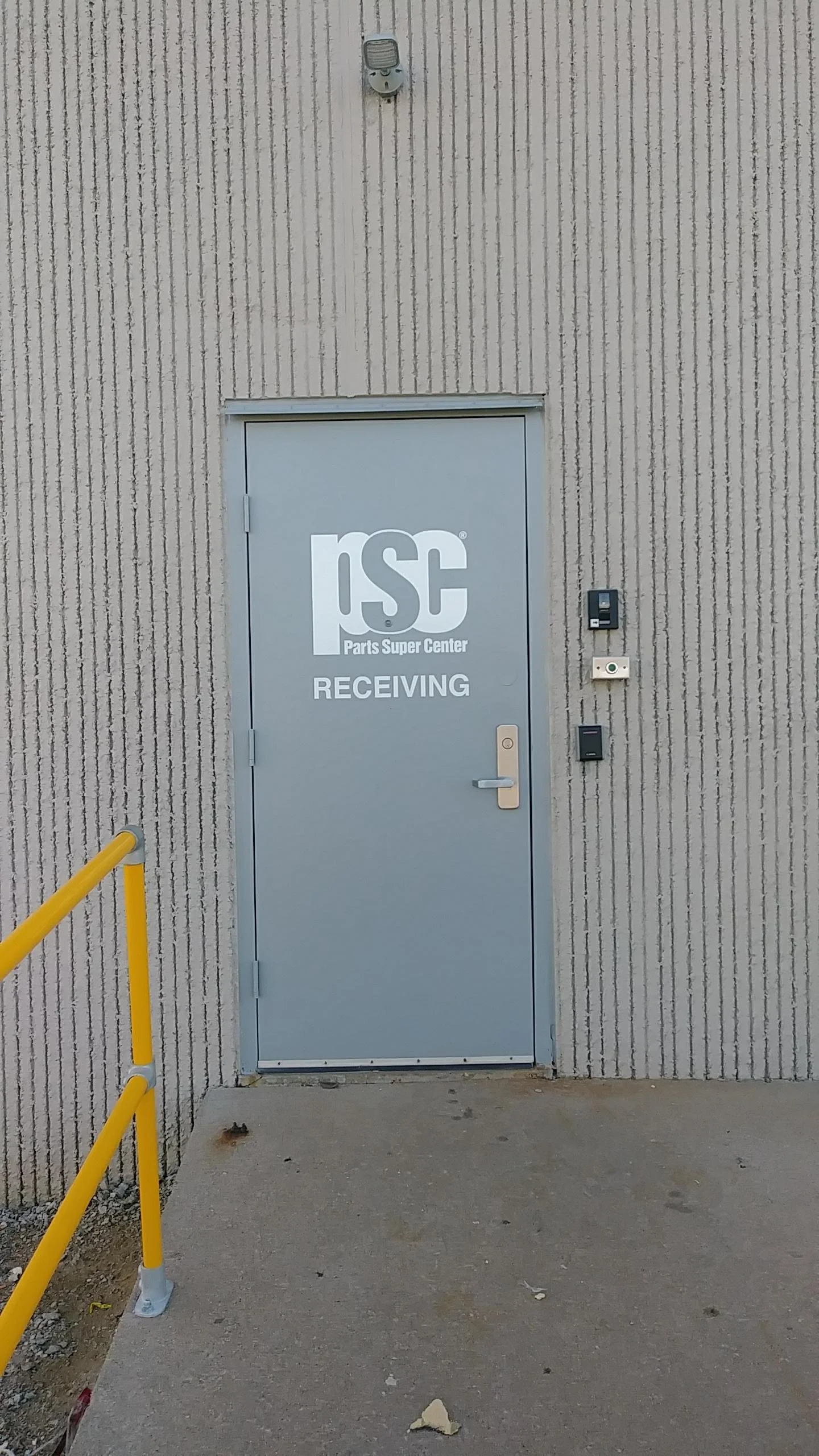 Gray door with 'ISC Parts Super Center RECEIVING' logo, beige textured wall, yellow safety rail, and security equipment on the wall.