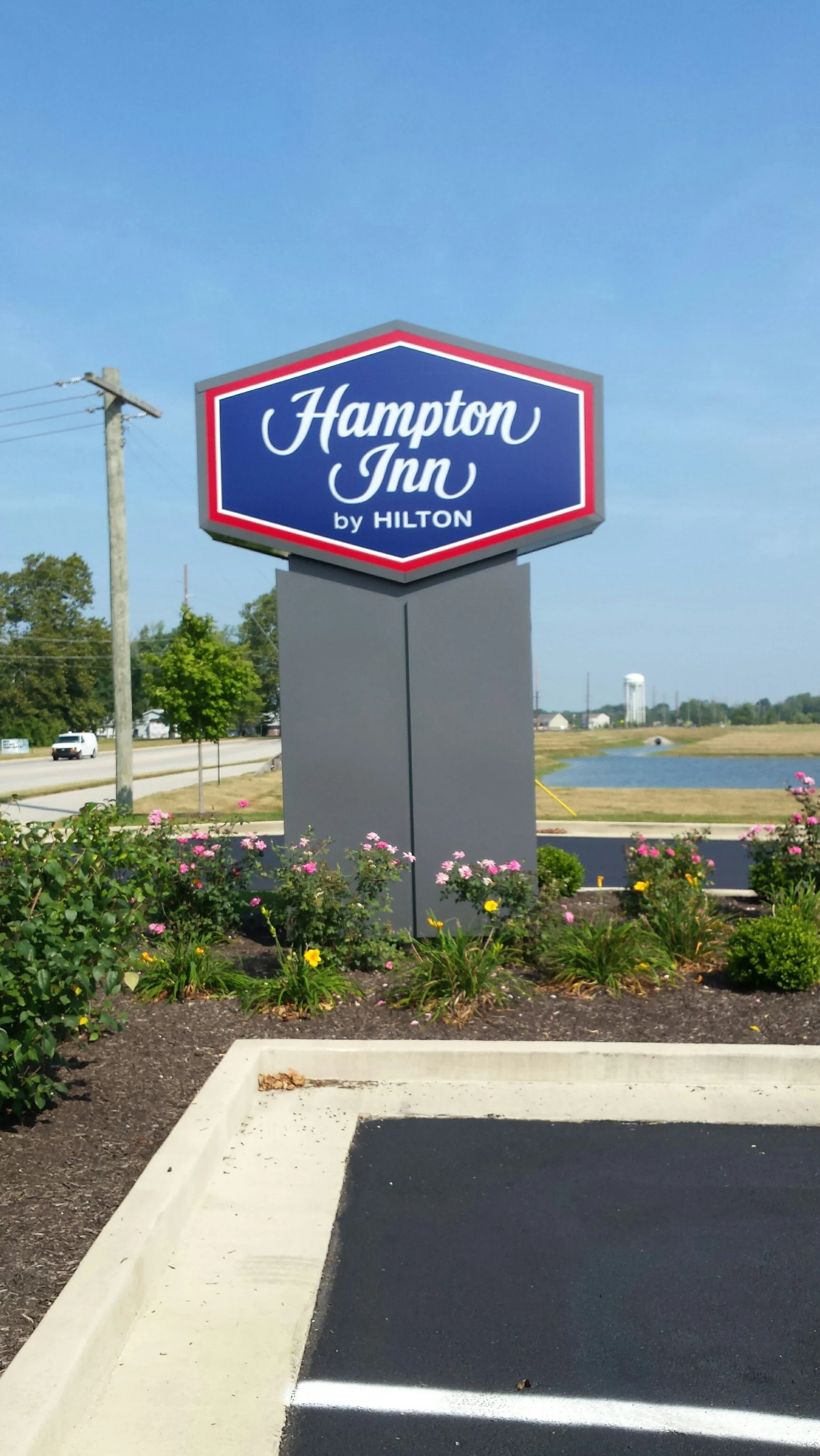Sign for Hampton Inn by Hilton with blooming flowers and parking lot in the foreground, open field with water and water towers in the background.