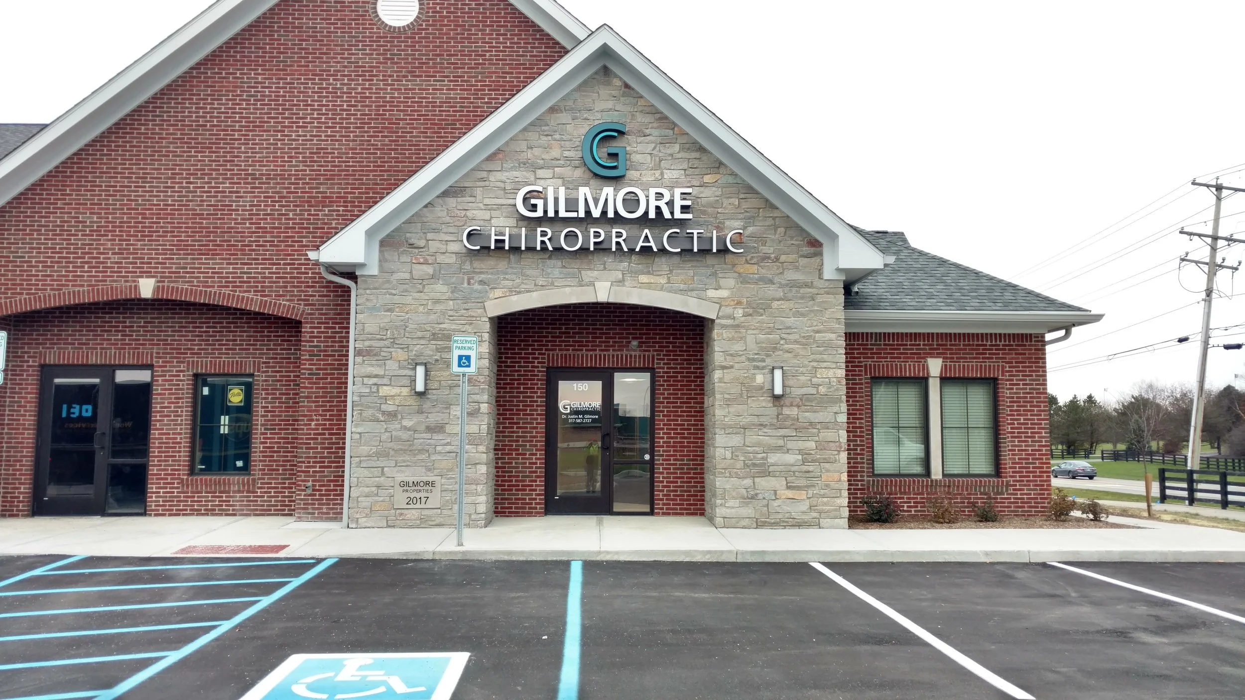 Front view of Gilmore Chiropractic building with red brick and stone facade, parking lot with handicap spaces, and power lines in the background.