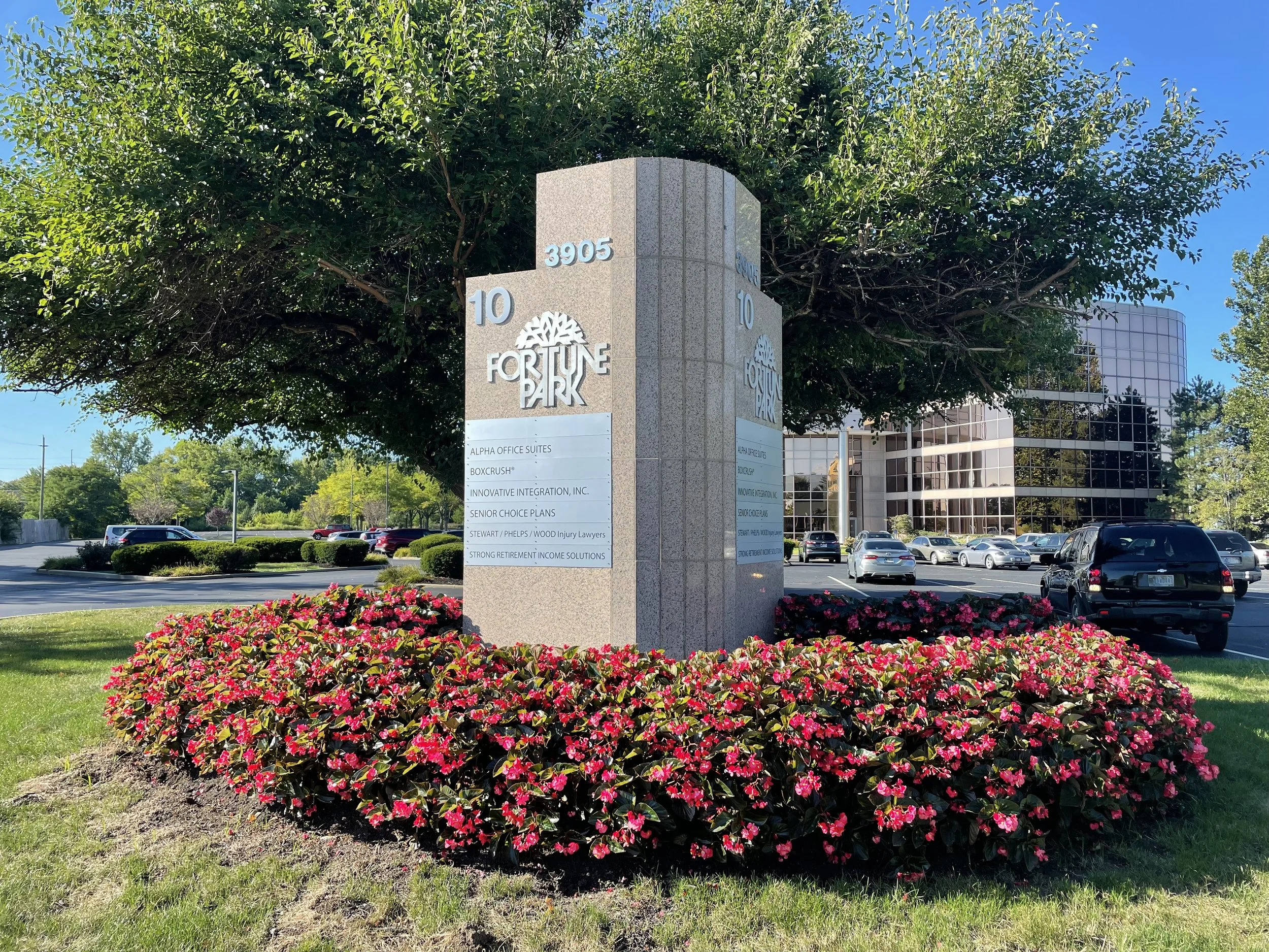 Sign at Fortune Park with businesses listed, surrounded by pink flowers, trees, and parking lot with cars in the background.