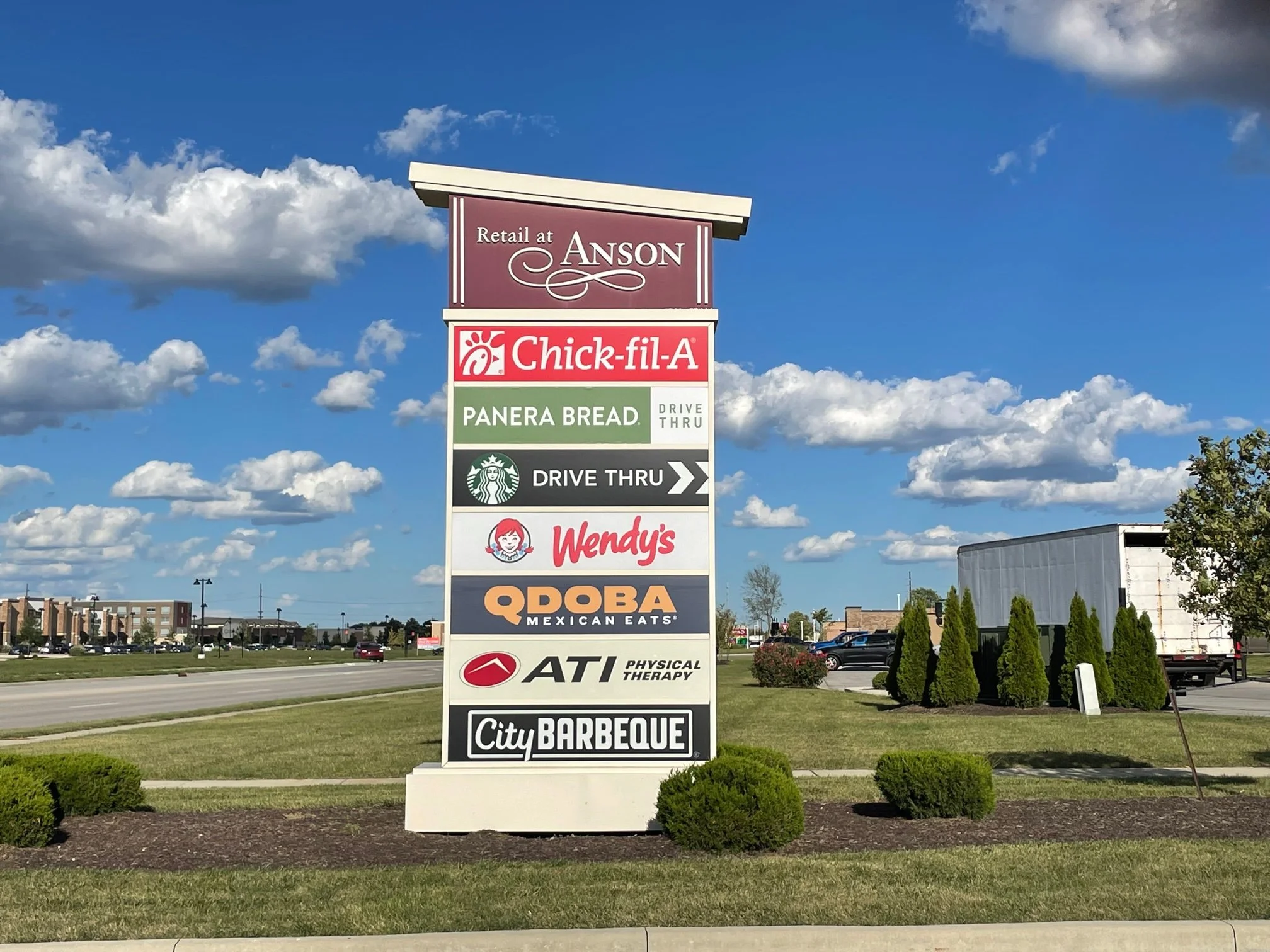 A large outdoor directory sign with multiple business logos, including Chick-fil-A, Wendy's, Taco Bell, ATI Physical Therapy, City Barbeque, and others, positioned on a grassy area near a road with a partly cloudy sky overhead.