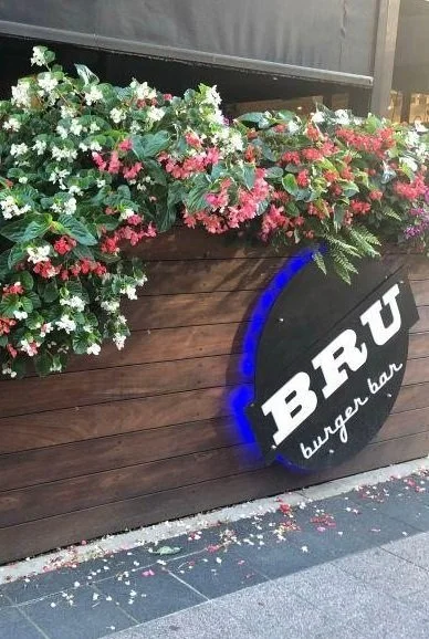 A wooden planter box filled with white, pink, and red flowers decorated with green leaves. The planter has a black round sign on it with the words "BRU burger bar" and a blue neon light accent. Flower petals are scattered on the ground below.