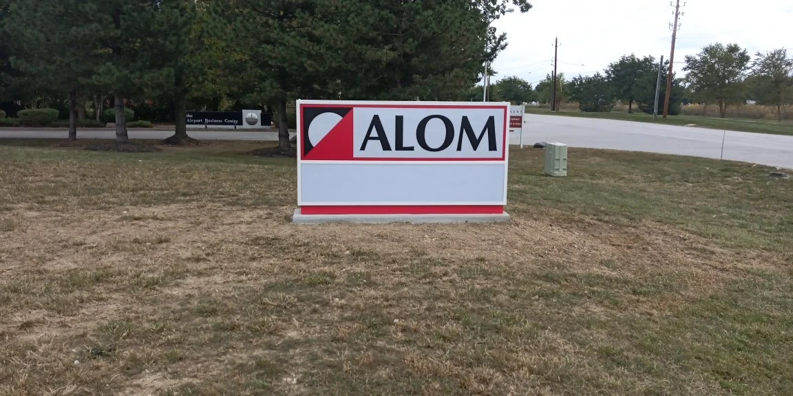 A sign with the word 'ALOM' on it, partially obscured by a shadow, on a grassy area near a road with trees and utility boxes in the background.
