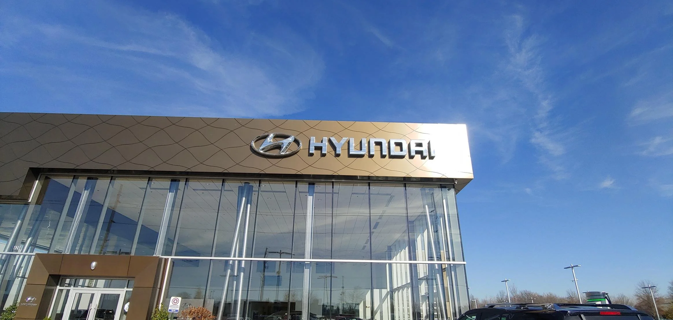 Exterior view of a Hyundai dealership building with glass windows and the Hyundai logo on the upper facade, under a partly cloudy blue sky.