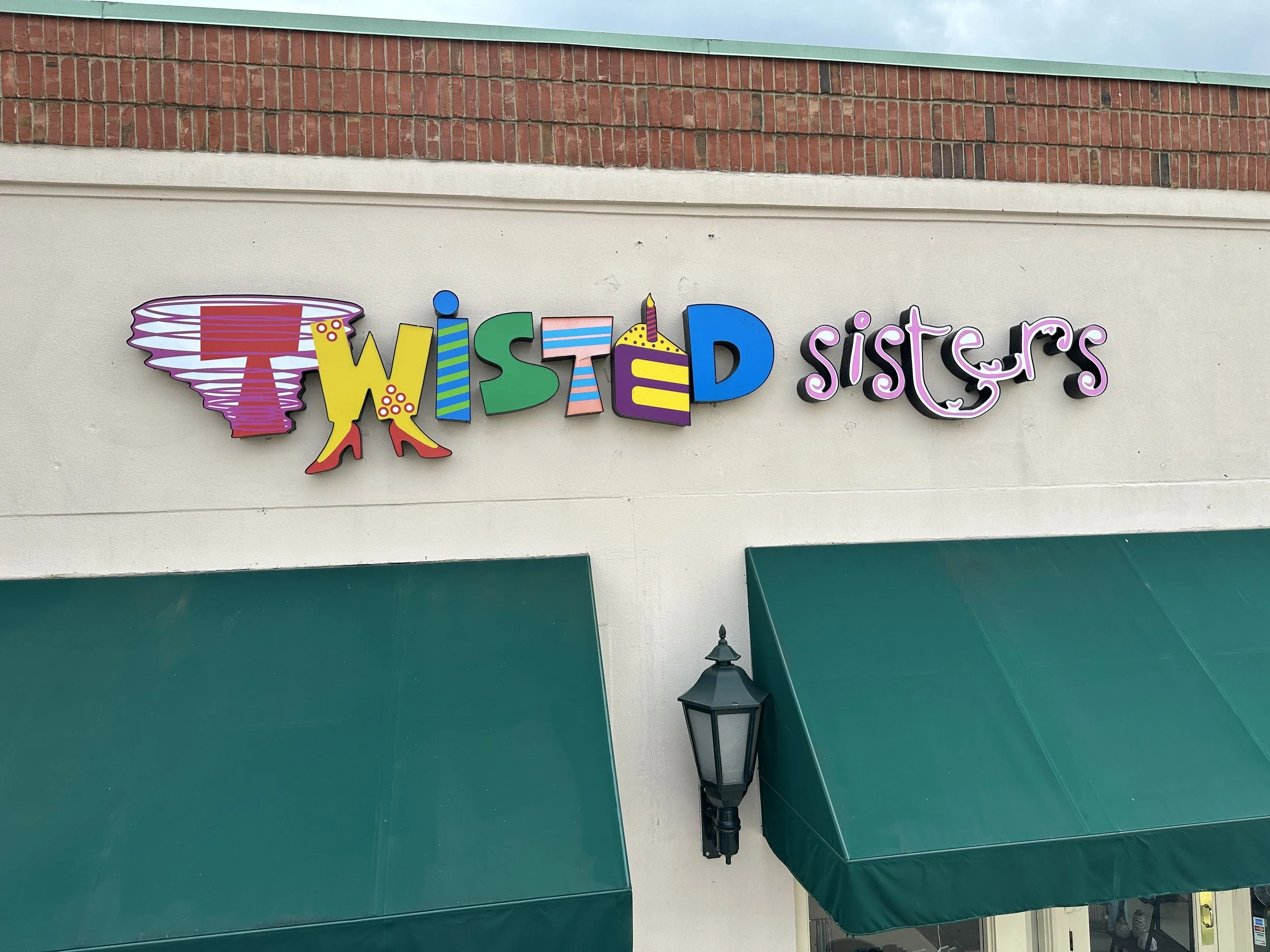 Sign for Twisted Sisters with colorful, playful lettering and a spinning tornado graphic on the left, above storefront with green awnings and a black lamp post.