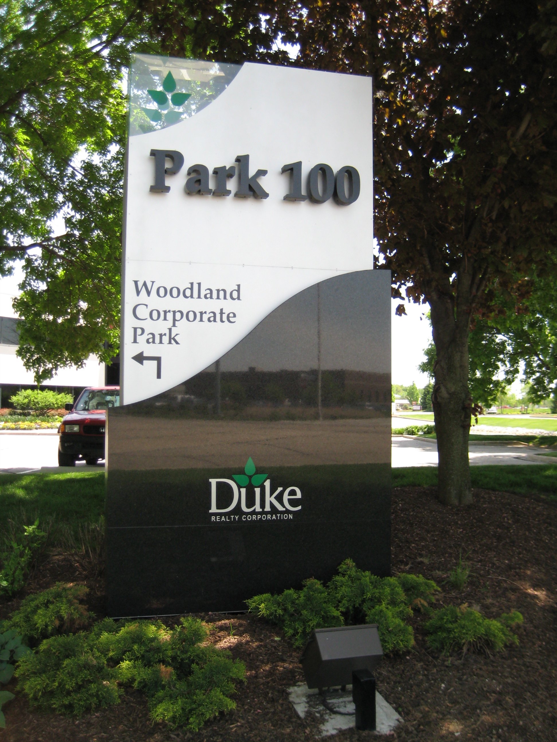 Sign for Park 100 in front of trees, with a building, a red vehicle, and a landscape background, featuring the Duke Realty Corporation logo and a direction arrow for Woodland Corporate Park.