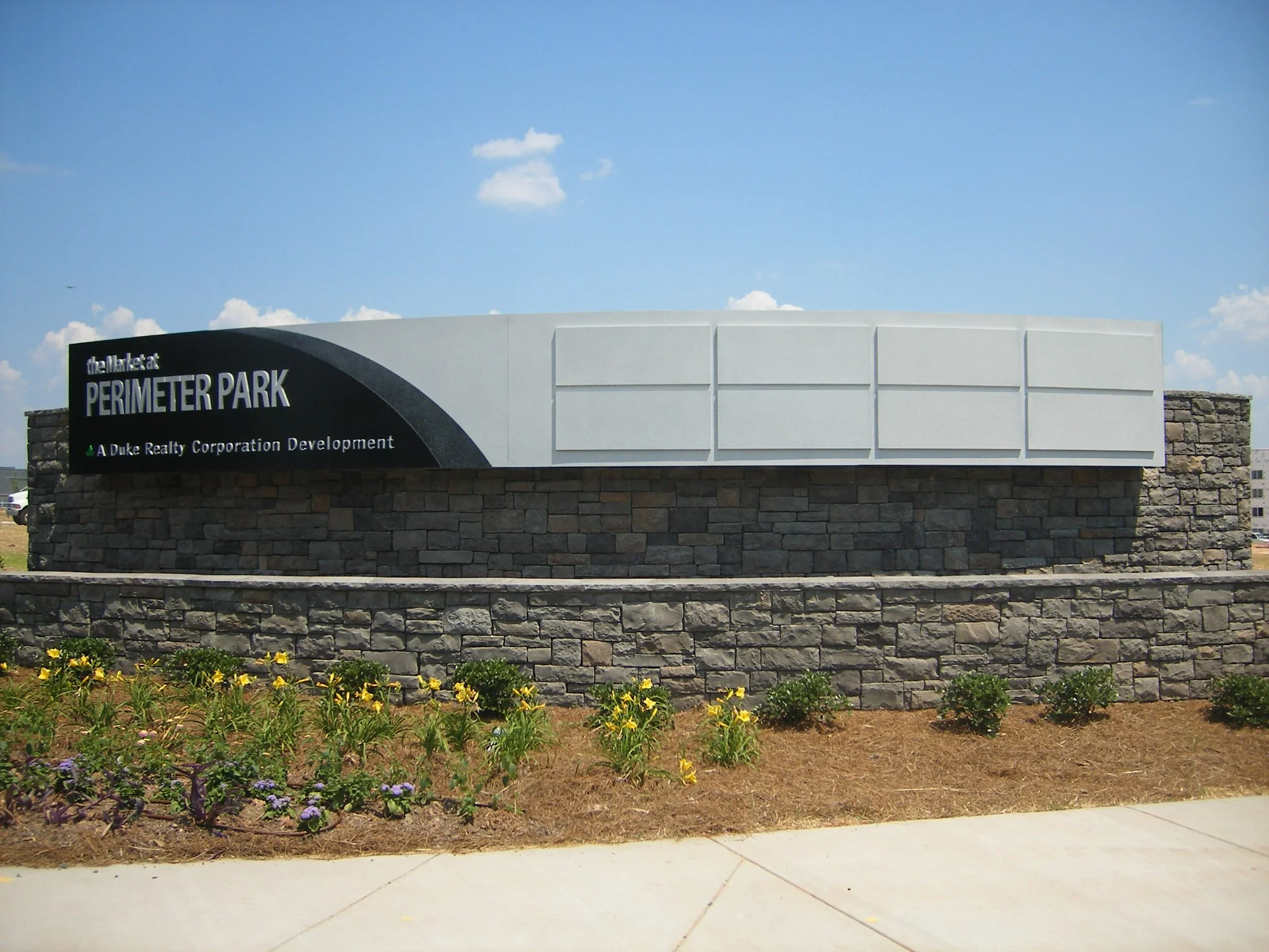 Signboard for Perimeter Park development, with a stone base, white upper part, and black background with white text, situated in a landscaped area with flowers and greenery.