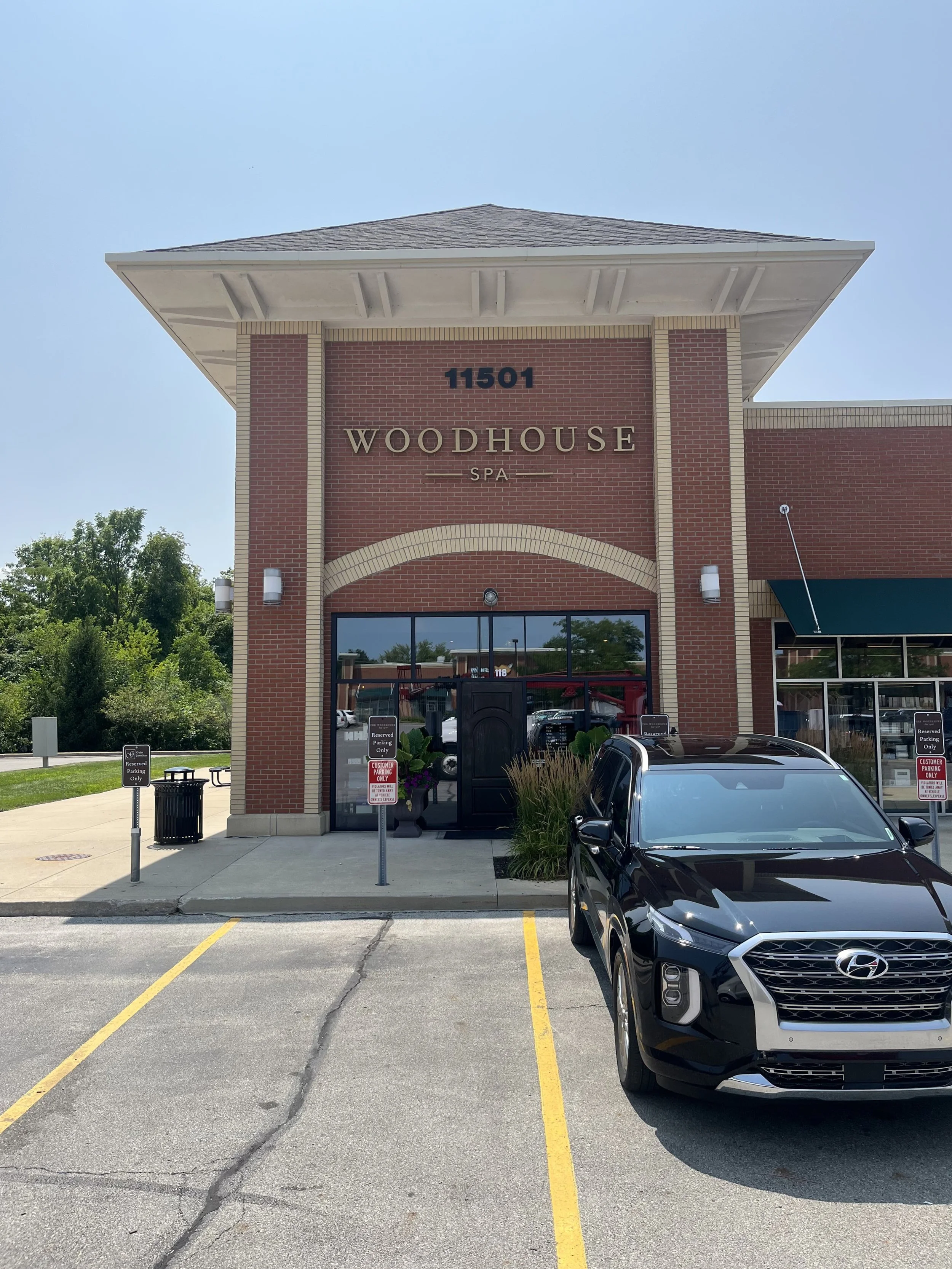 Brick building with a sign that reads 'Woodhouse Spa' and the address 11501 above the entrance. A black Hyundai car is parked in front, and there are reserved parking signs nearby.