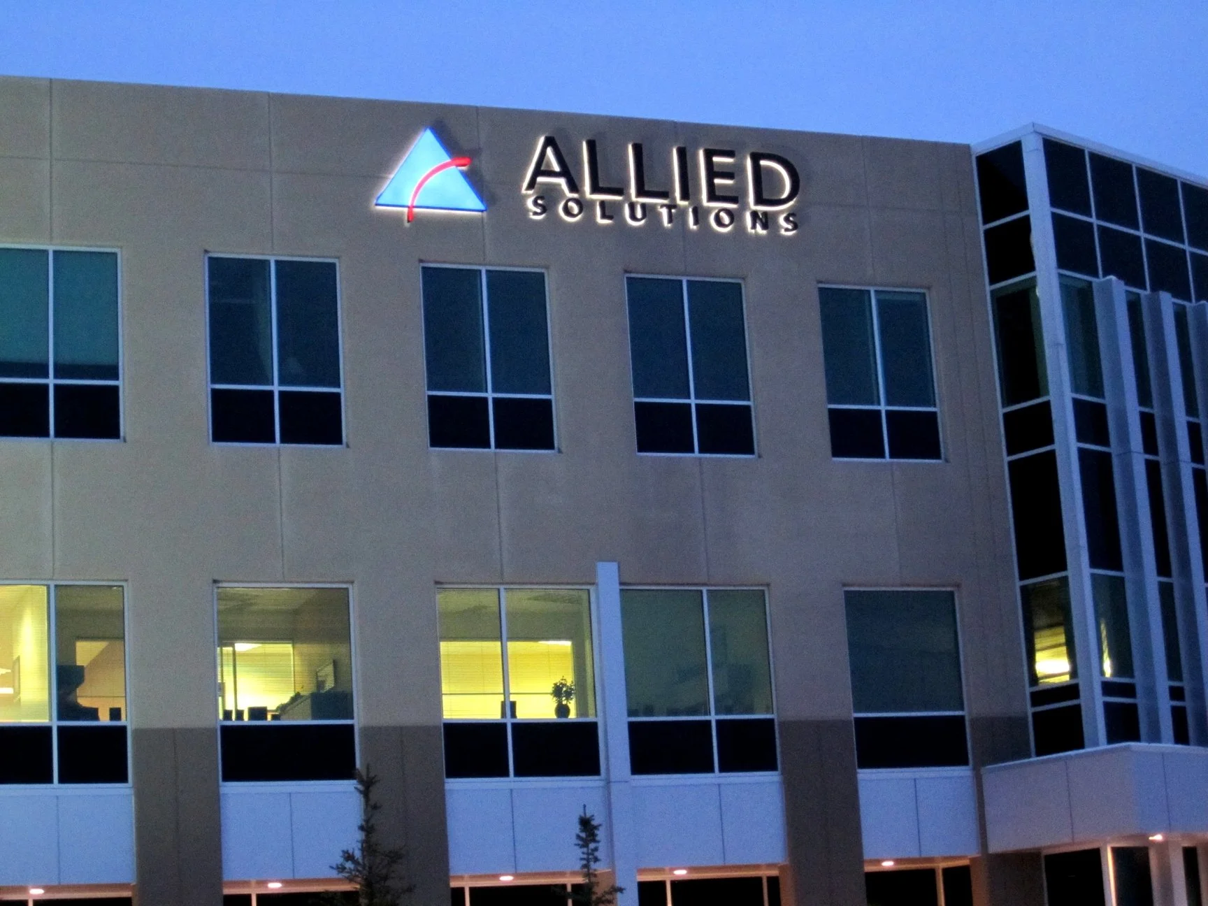 Exterior of a modern office building with a sign that reads 'ALLIED SOLUTIONS' and a logo featuring a stylized triangle with red and blue accents, during dusk or evening.