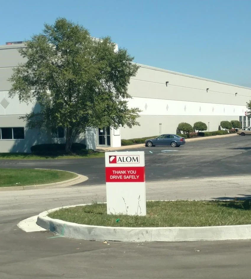 A parking lot with a large tree and a building in the background. In the foreground, a white sign with red and black text reads "Alom" with a logo, and below it, "Thank you. Drive safely."