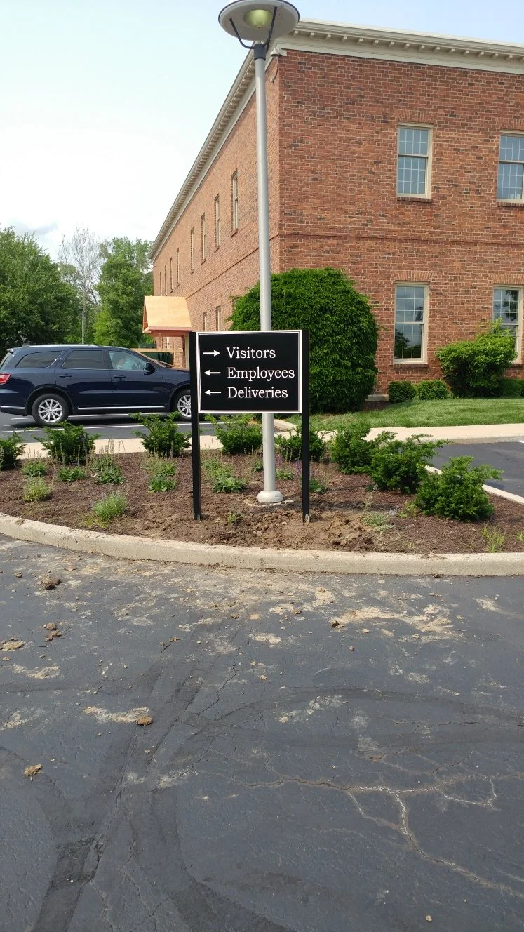 A black sign with white arrows directing visitors to the right, employees to the left, and deliveries to the left, in a parking lot near a brick building with a landscaped area.