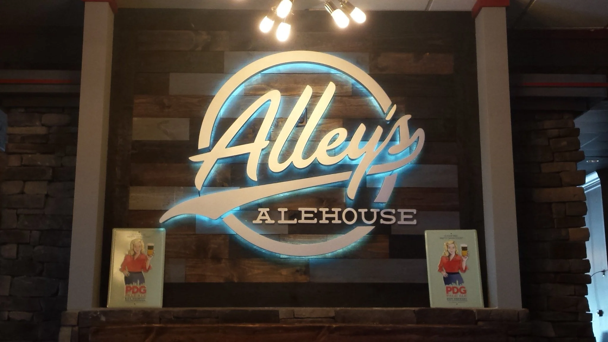Illuminated sign for Alley’s Alehouse on a wooden wall with two posters of a woman holding a beer at the bottom.