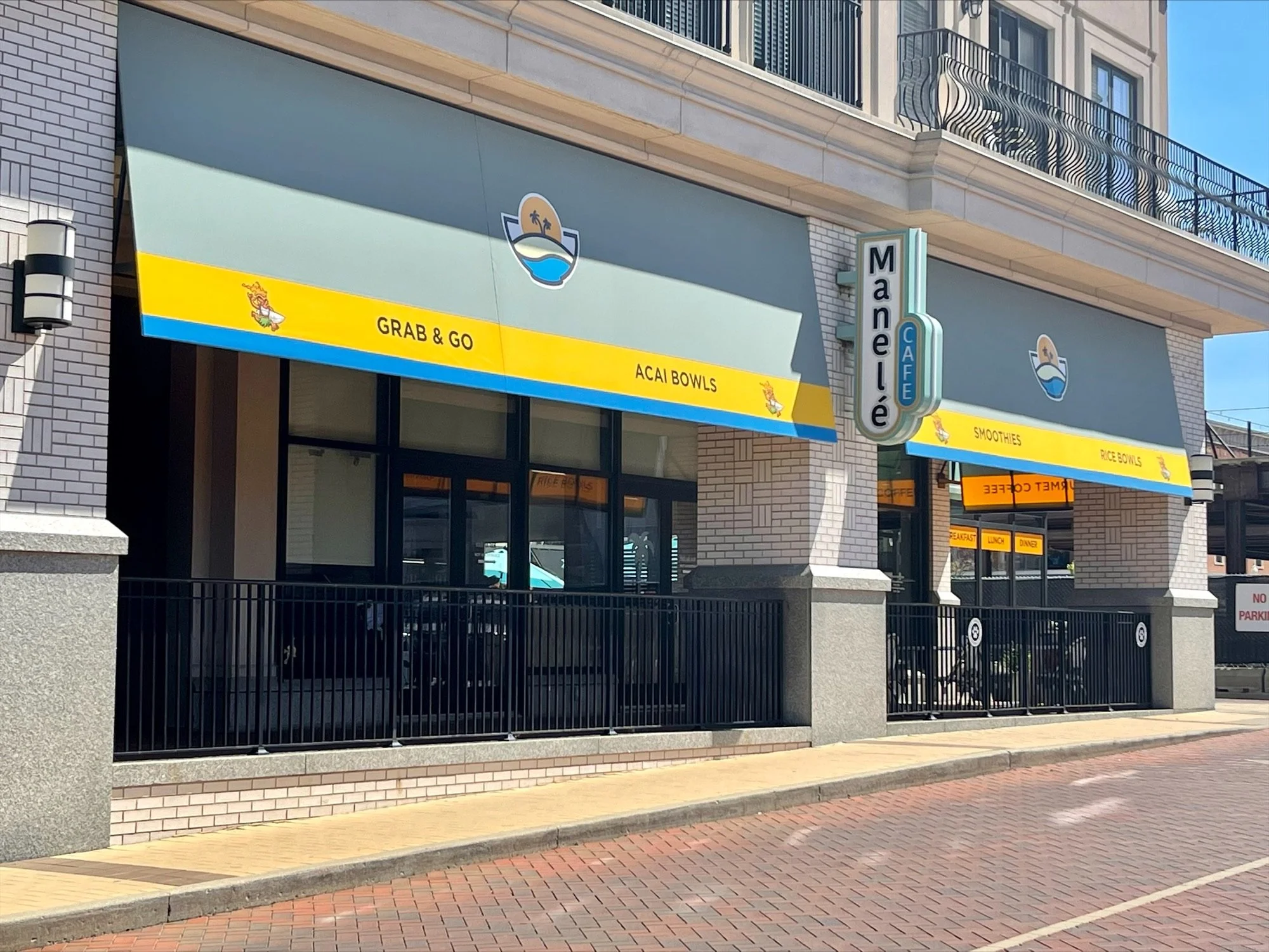 Exterior of Maneilé Café with blue awnings advertising grab-and-go food, acai bowls, smoothies, rice bowls, and breakfast, lunch, dinner options. The café has a neon sign and outdoor seating area with black railing.