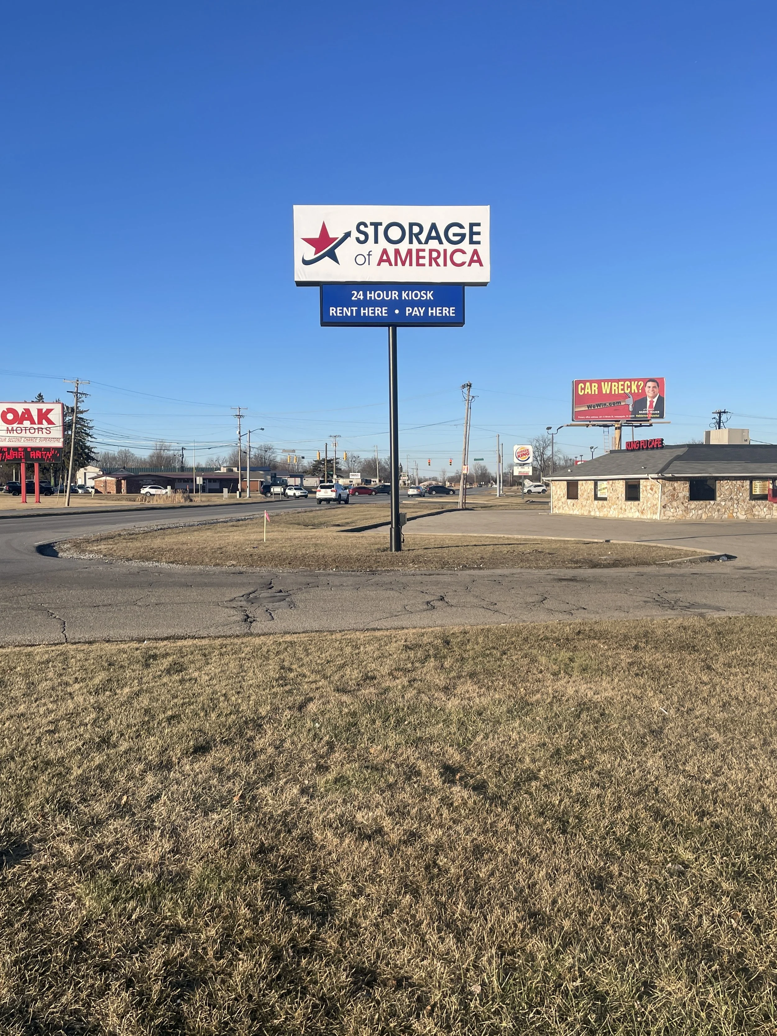 A large sign for Storage of America in a parking lot, with a blue sky background. The sign indicates 24-hour kiosk for renting and paying.