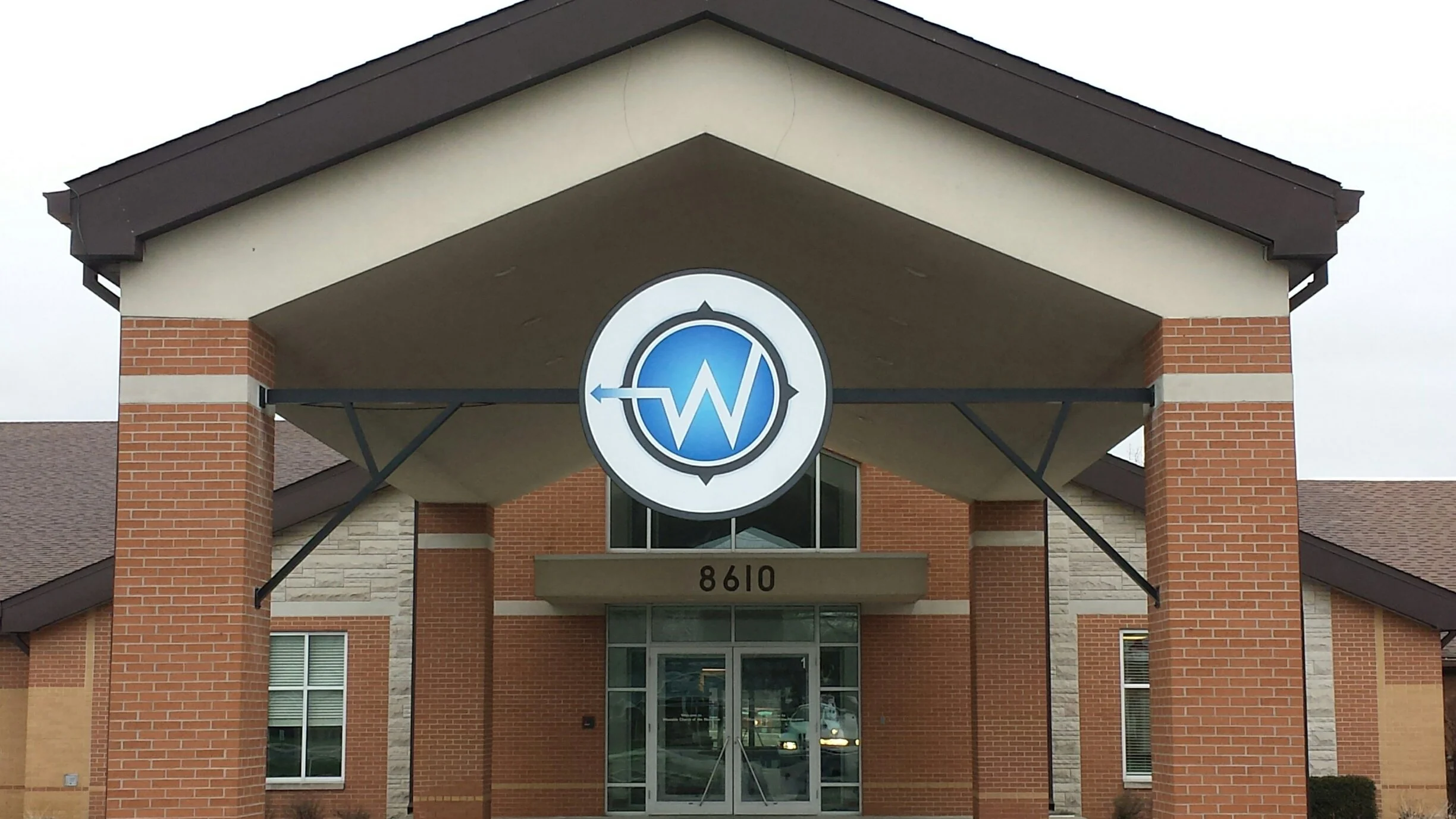Front view of a brick building with the number 8610 above a glass door entrance, featuring a round logo with a blue circle, a black compass-like border, and a white zigzag line inside a blue background, mounted under a porch roof.