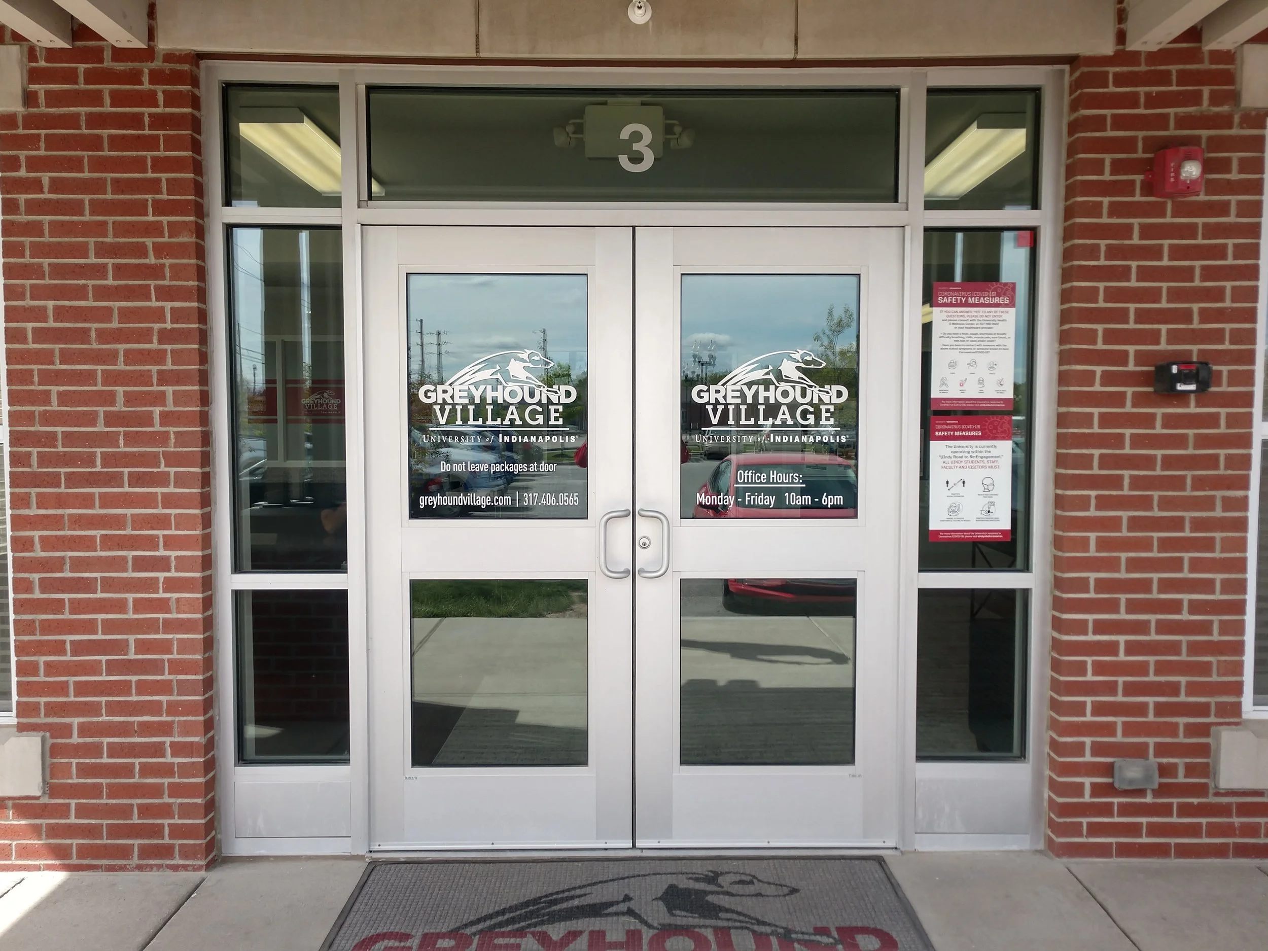 Double glass doors with the Greyhound Village logo and text, located in a brick building, with office hours and safety measures posted.