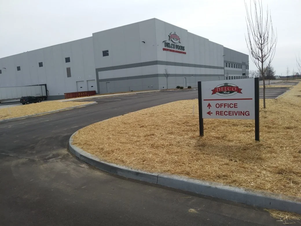 Large industrial building with signs indicating office and receiving areas, paved driveway, and a few leafless trees in a mostly empty parking lot and landscape.