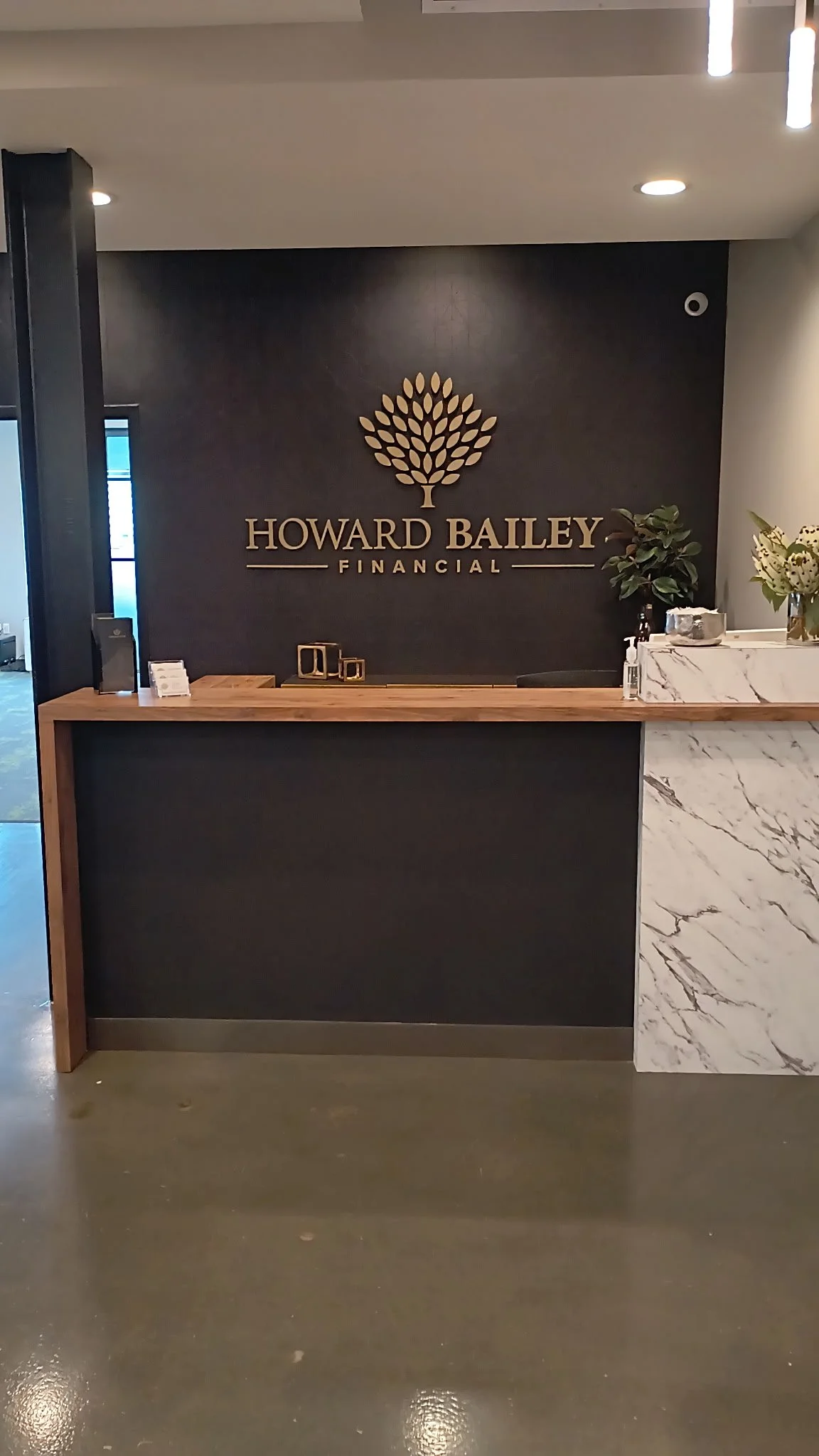 Reception desk with a black wall behind displaying the Howard Bailey Financial logo and name, decorated with a gold tree emblem, a plant, and a marble and wood counter.