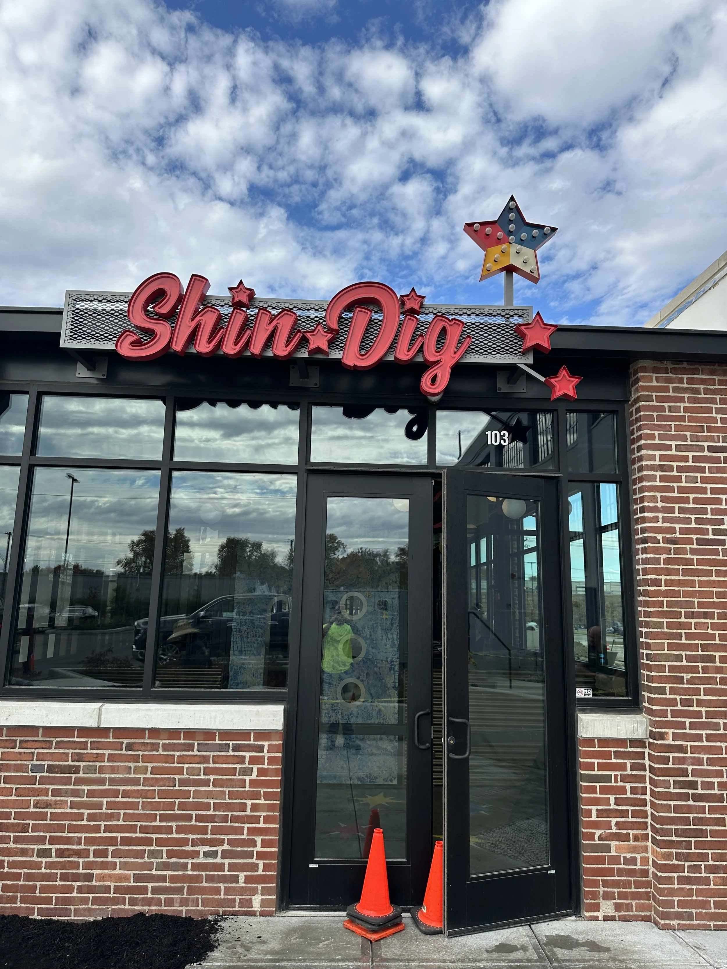 Exterior of a restaurant named Shin Dig with red neon sign, brick walls, large glass windows, and a decorative star on the roof. Two orange cones are placed by the entrance.