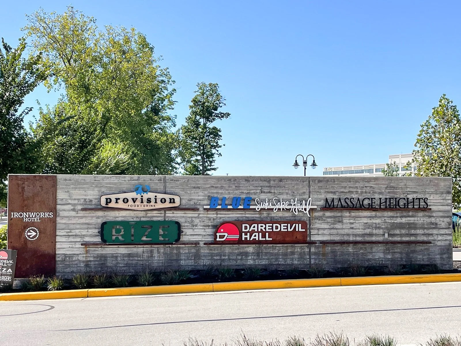 A large cement wall with various business signs, including Provision, Blue Sushi Sushi Grill, Massage Heights, Rize, Daredevil Hall, and Ironworks Hotel, with trees and a clear blue sky in the background.