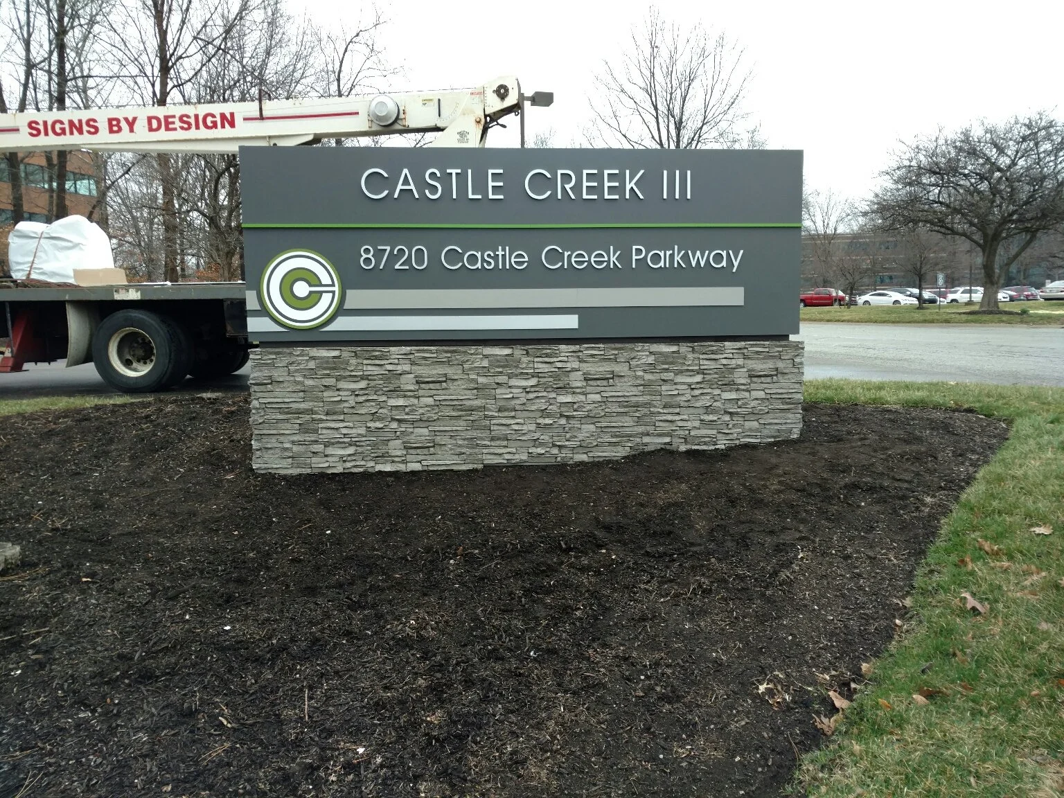 Sign for Castle Creek III at 8720 Castle Creek Parkway, with a stone base and gray background, partially obscured construction truck in the background, and a parking lot with cars and leafless trees.