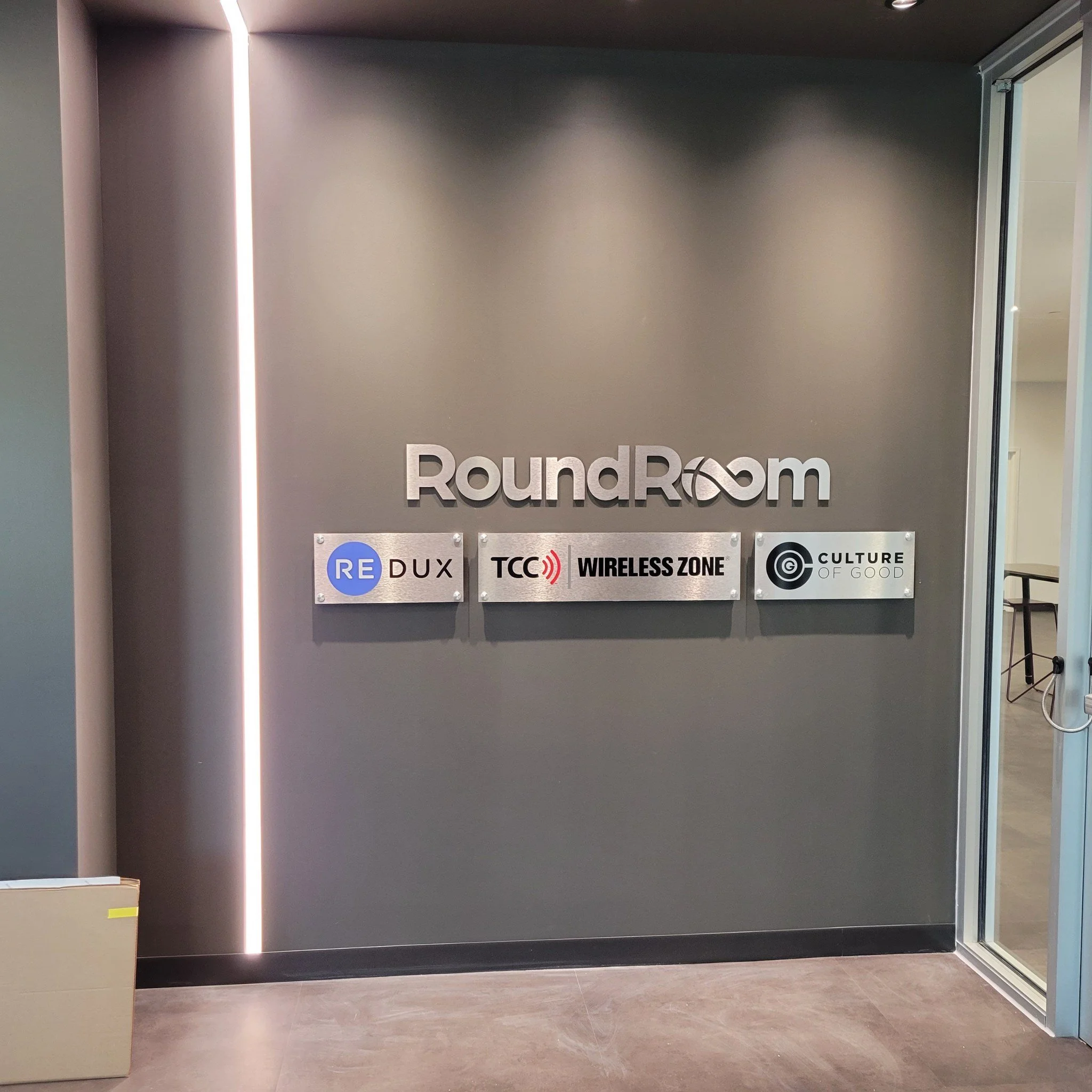 Sign reading 'RoundRoom' with additional sponsor logos below, including RE DUX, TCC Wireless Zone, and Culture of Good, on a dark gray wall.