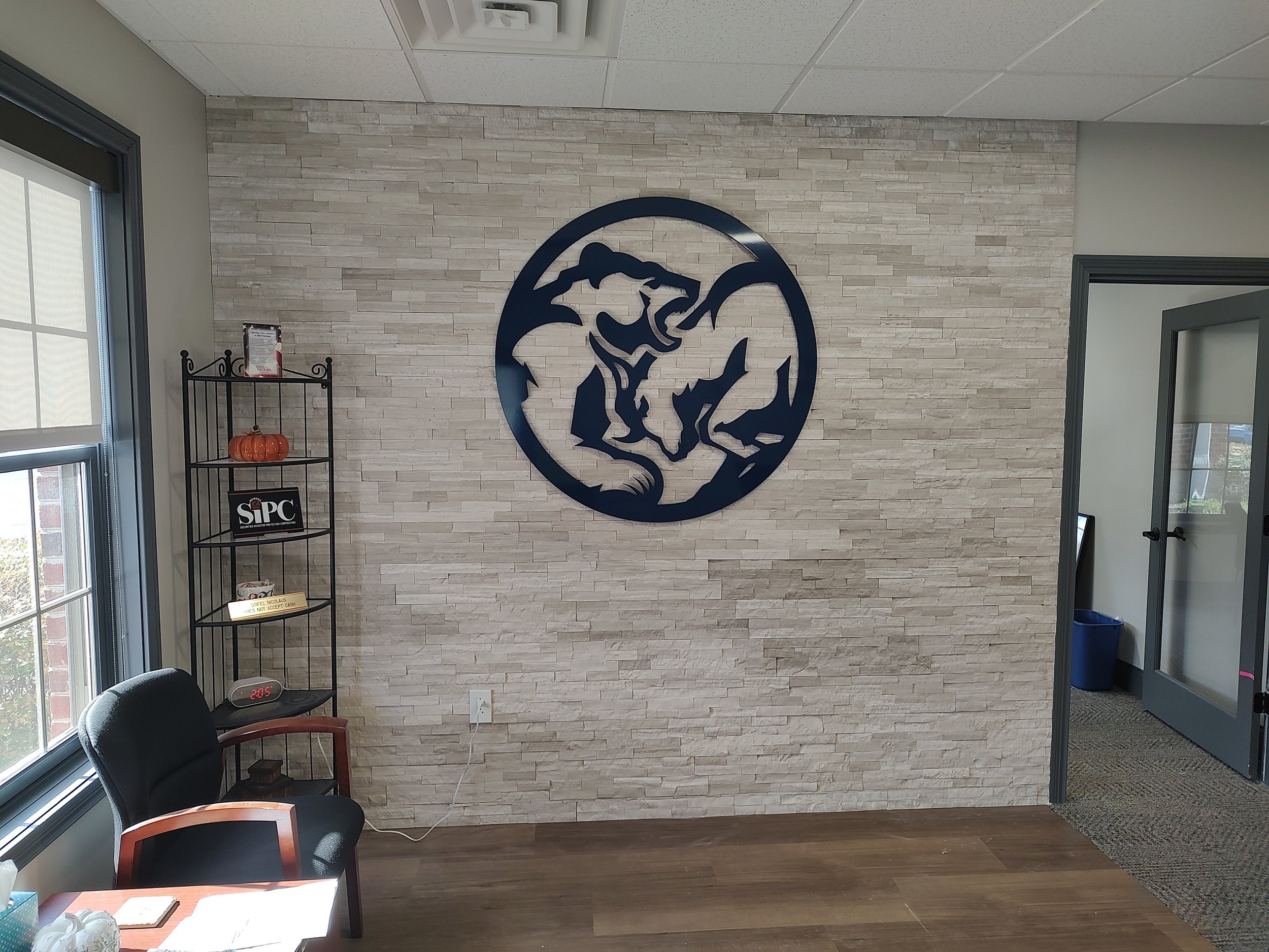 Interior of an office with a beige brick accent wall featuring a black circular logo of a howling wolf. There is a black chair and a small shelving unit with decorative items, including a pumpkin and a sign that says SIPC. A window on the left side b