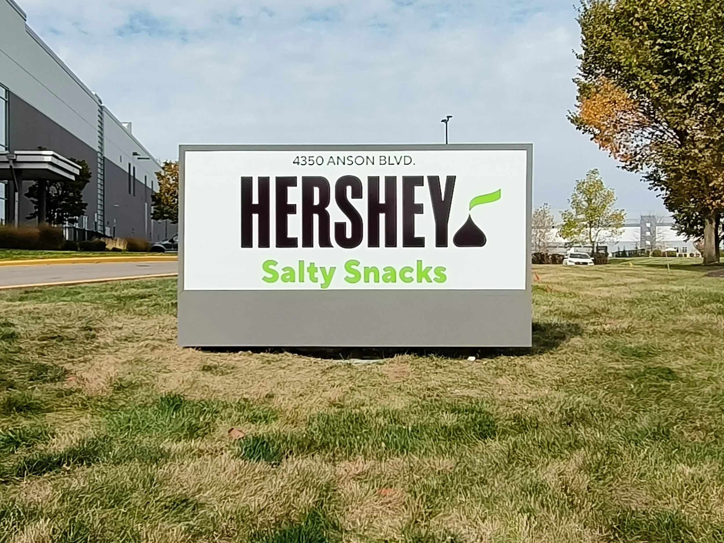 Sign for Hershey Salty Snacks located at 4350 Anson Blvd, outdoors on a grassy area with trees and a building in the background.