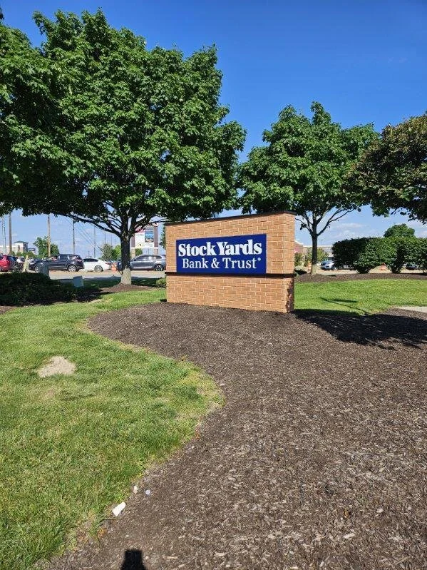 A sign reading "Stock Yards Bank & Trust" in front of three large green trees and a parking lot with cars, under a bright blue sky.