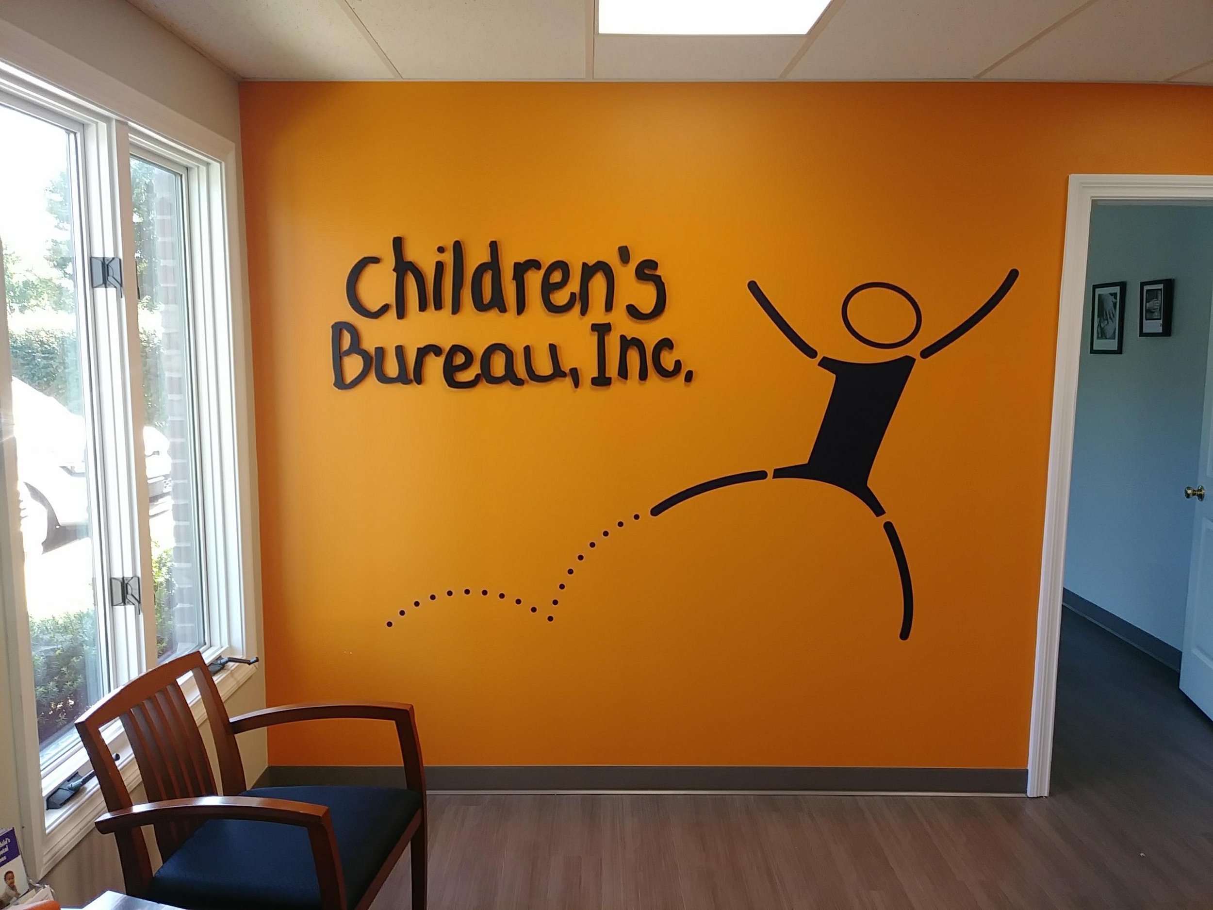 Interior of a children's bureau with a bright orange wall, black text reading 'children's Bureau, Inc.', and a graphic of a joyful stick figure jumping.