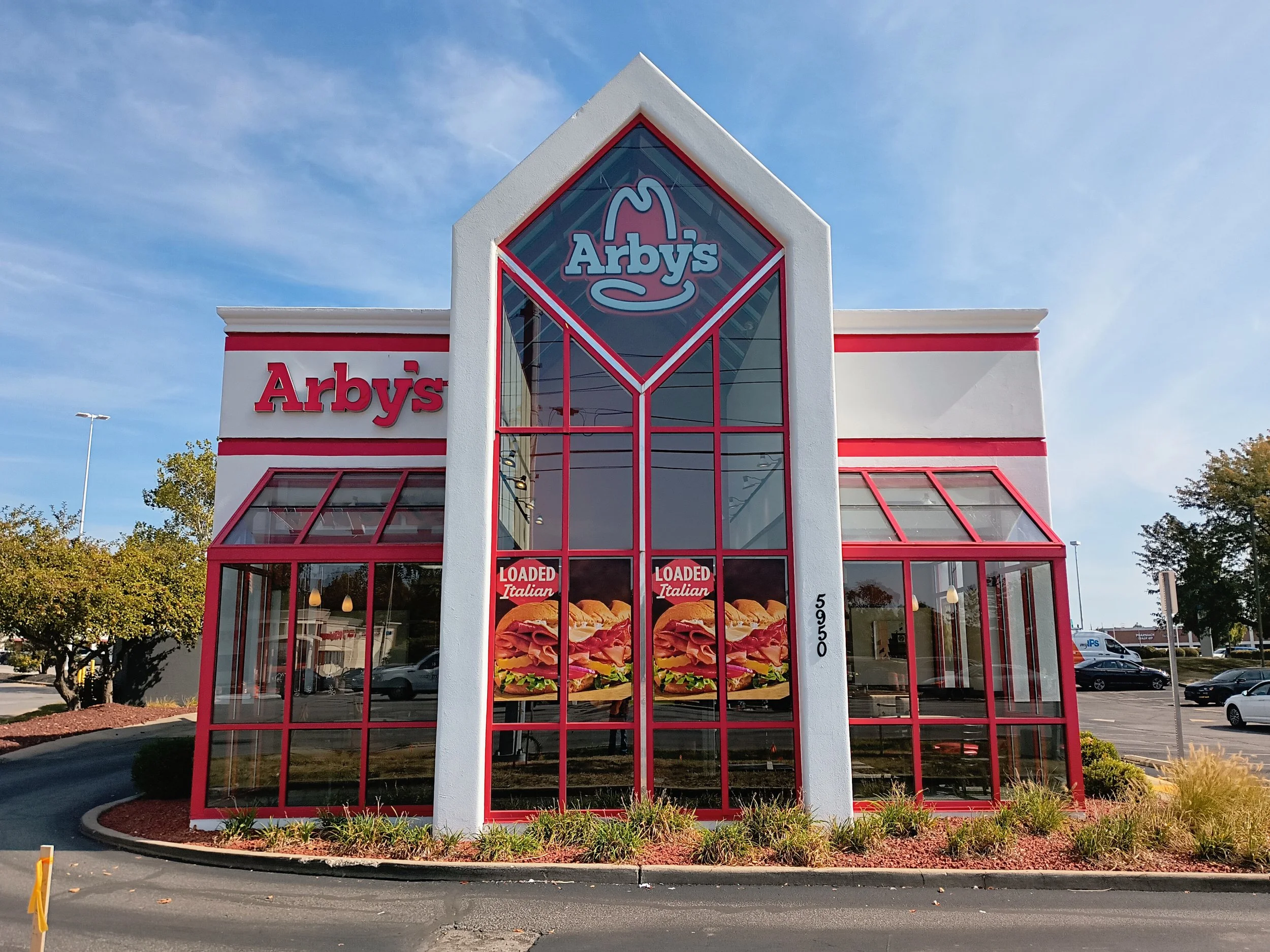 Front view of an Arby's fast food restaurant with large glass windows, red frame accents, and a prominent arched center section with the Arby's logo at the top. Posters of sandwiches are visible through the windows.
