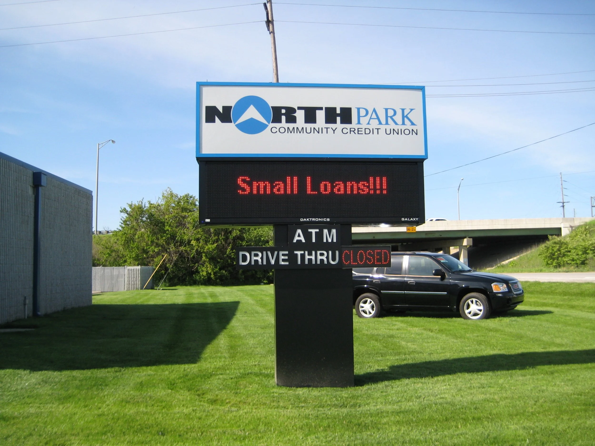 Sign for North Park Community Credit Union with electronic message board reading 'Small Loans!!!'. Below, a sign indicates ATMs and drive-thru are closed, with a black car parked on the grass nearby.