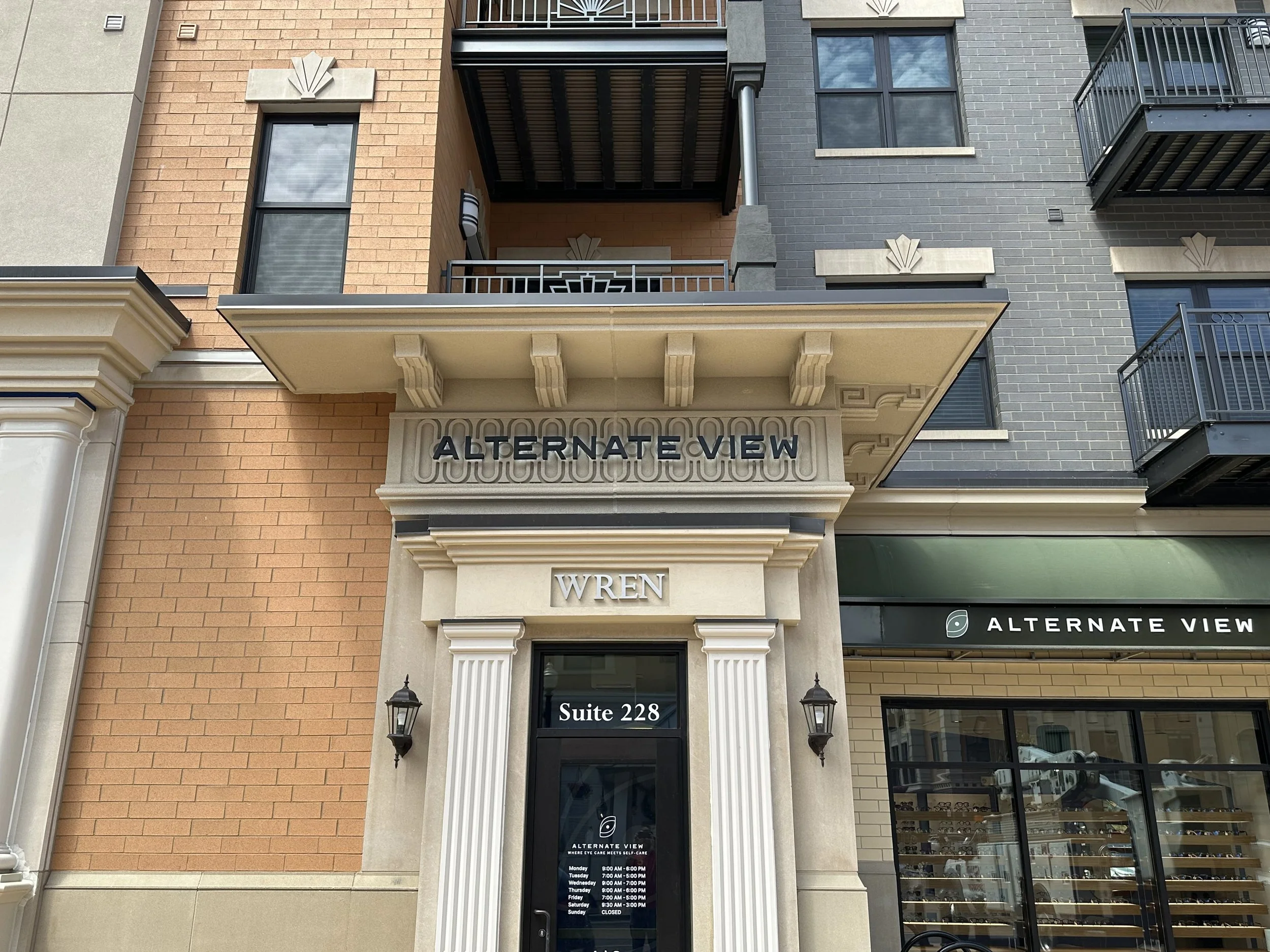 Facade of a building with a sign that reads "ALTERNATE VIEW" above a door marked "Suite 228". The building has brick and gray siding with balconies, large windows, and decorative architectural elements.