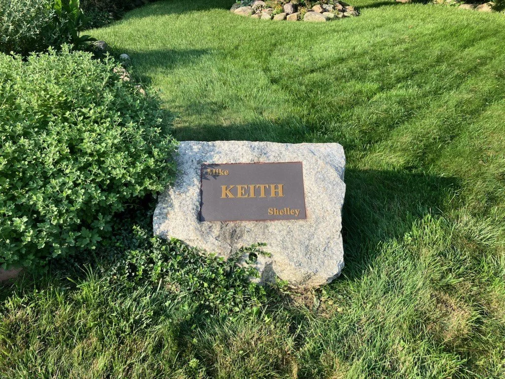 A stone with a metal plaque that reads 'Mike KEITH Shelley' placed on a lawn with trimmed grass and shrubs.
