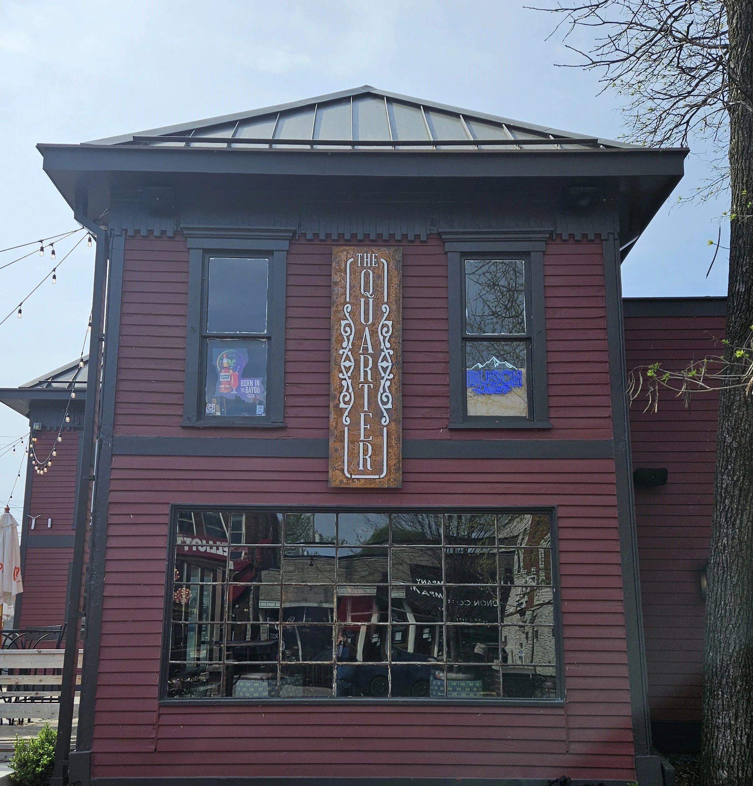 Two-story restaurant building with red siding and black trim, featuring a large vertical sign reading 'The Quarter' on the front. The upper windows have signs, one saying 'Born in Bayou' and another neon sign that reads 'Busch Light.' Reflective larg