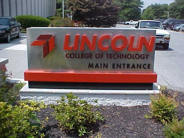 Sign for Lincoln College of Technology, main entrance, outdoor setting with parking lot and cars in the background.