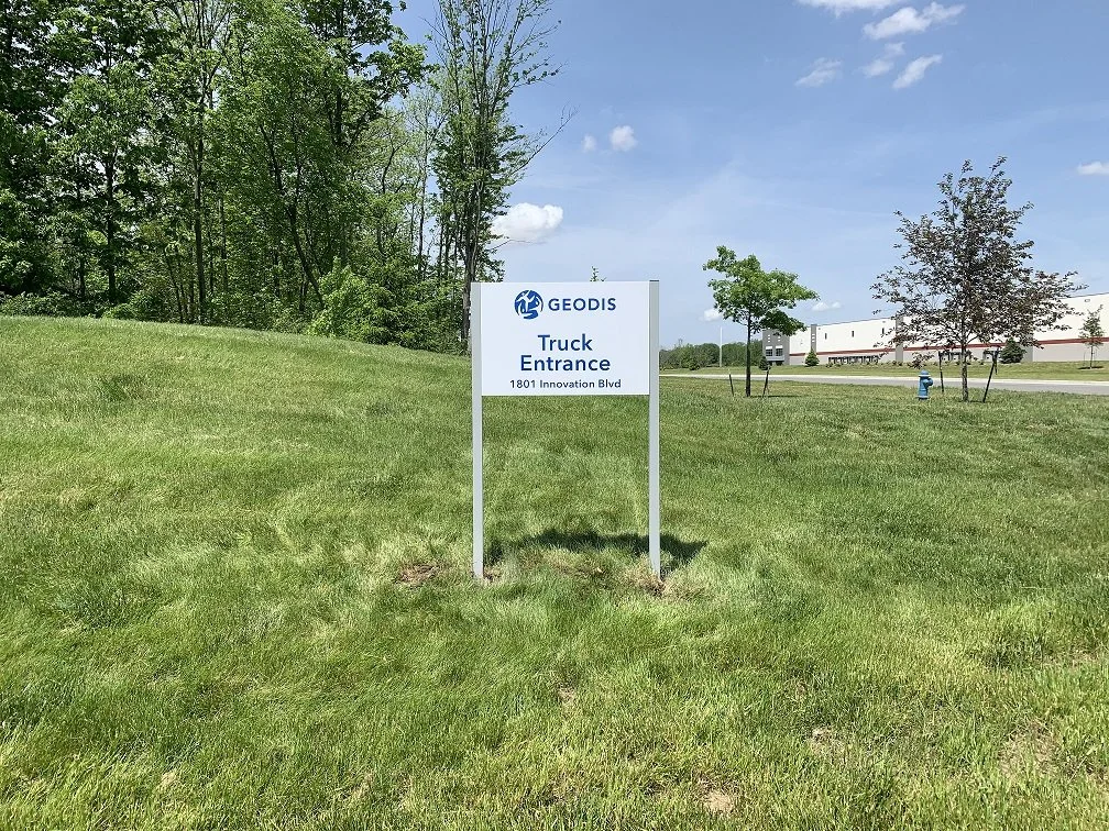 A sign reading 'GEODIS Truck Entrance 1801 Innovation Blvd' is placed on a grassy area near a road, with trees and a building in the background under a partly cloudy sky.