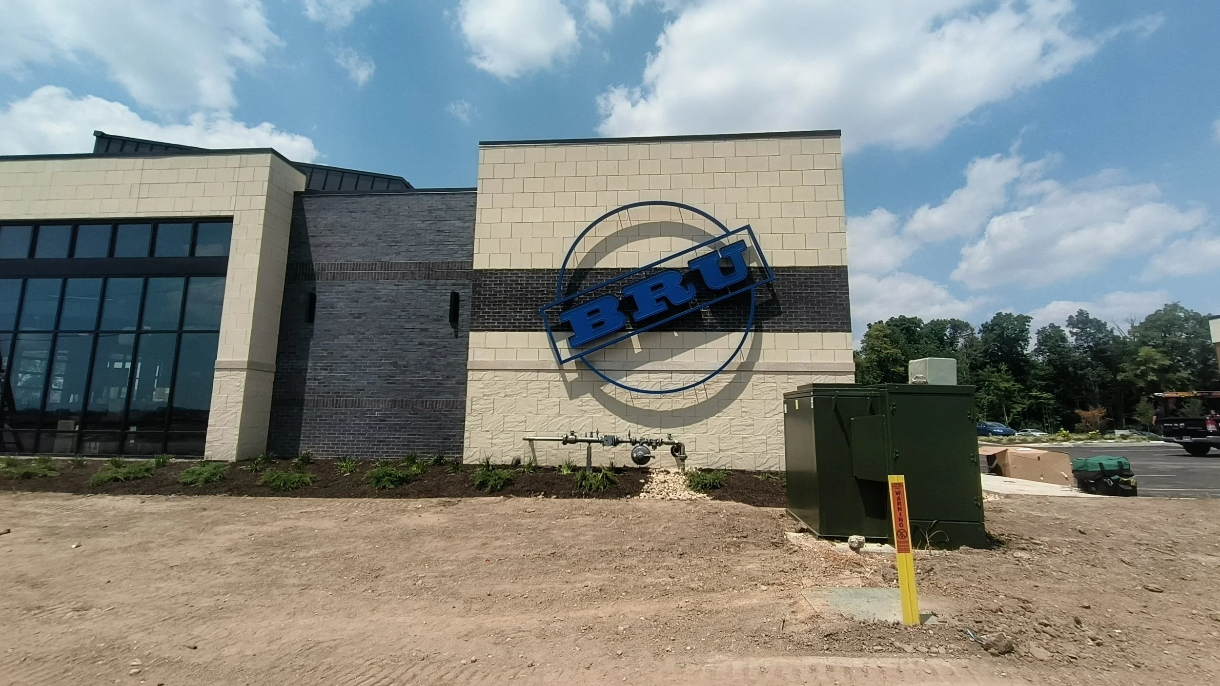Side view of a modern building with a large sign that reads 'BRU' on an exterior wall, with a partly cloudy sky and parking lot in the background.