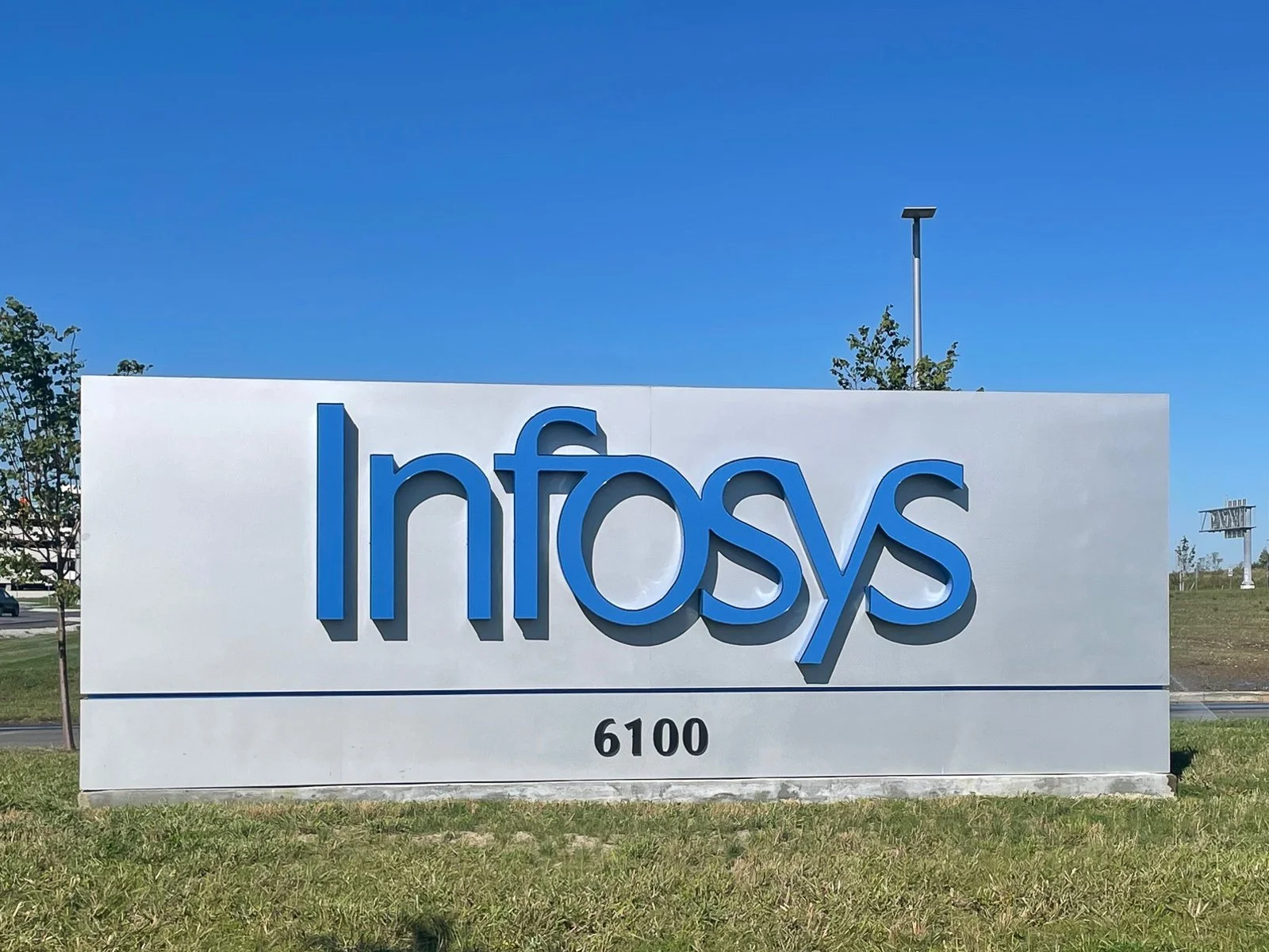 Large white sign with the name 'Inmosy' in blue letters and the number 6100 below it, outdoors on grass with trees and blue sky in the background.