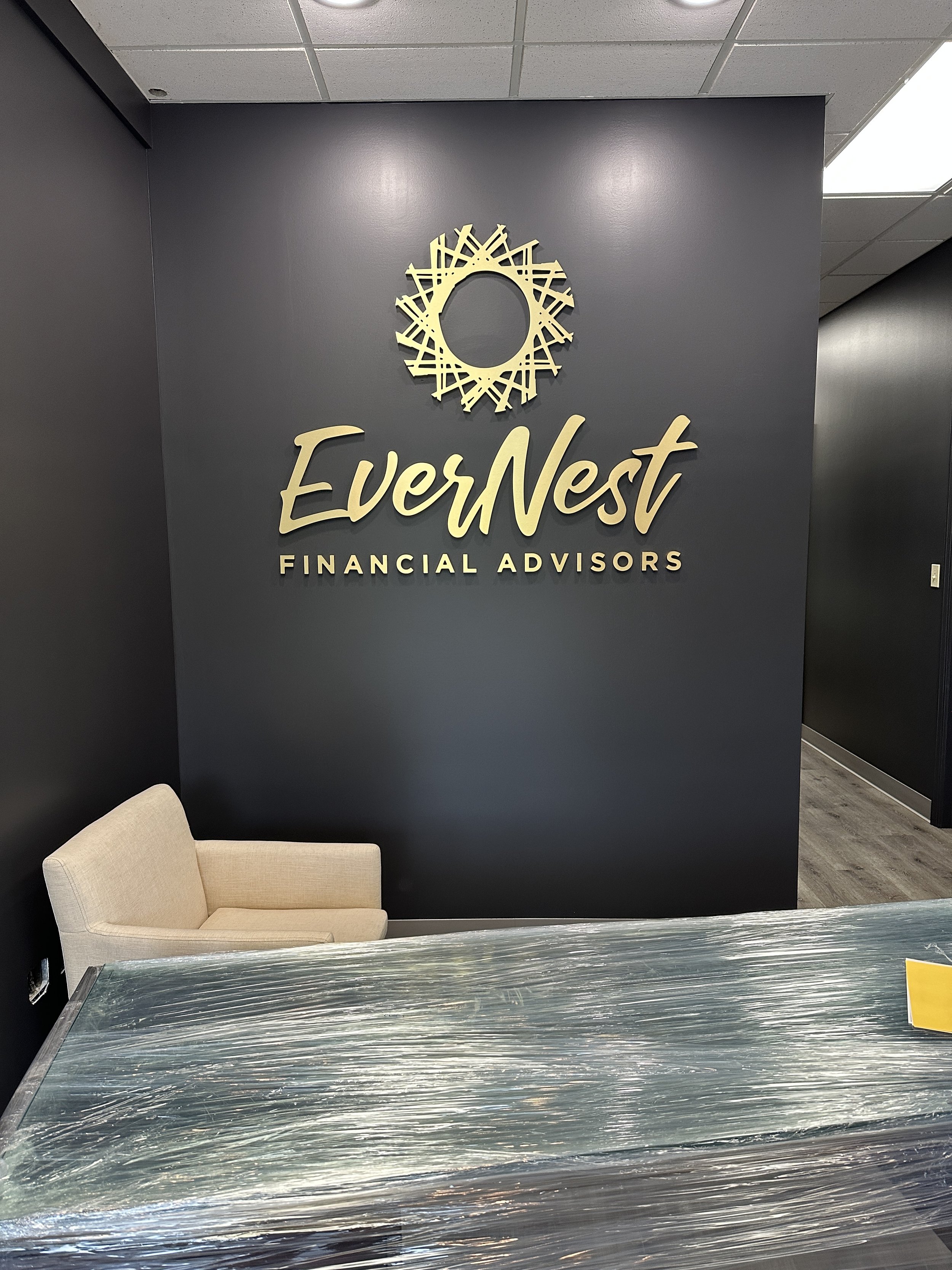 Reception area with a dark wall displaying the logo for EverNest Financial Advisors, featuring a circular geometric design above the business name. There is a beige chair on the left and a glass-covered table in the foreground.