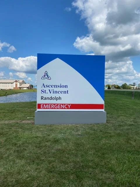 Sign for Ascension St. Vincent in Randolph with a blue and white design, and an emergency section highlighted in red, placed on a green grass field under a partly cloudy sky.