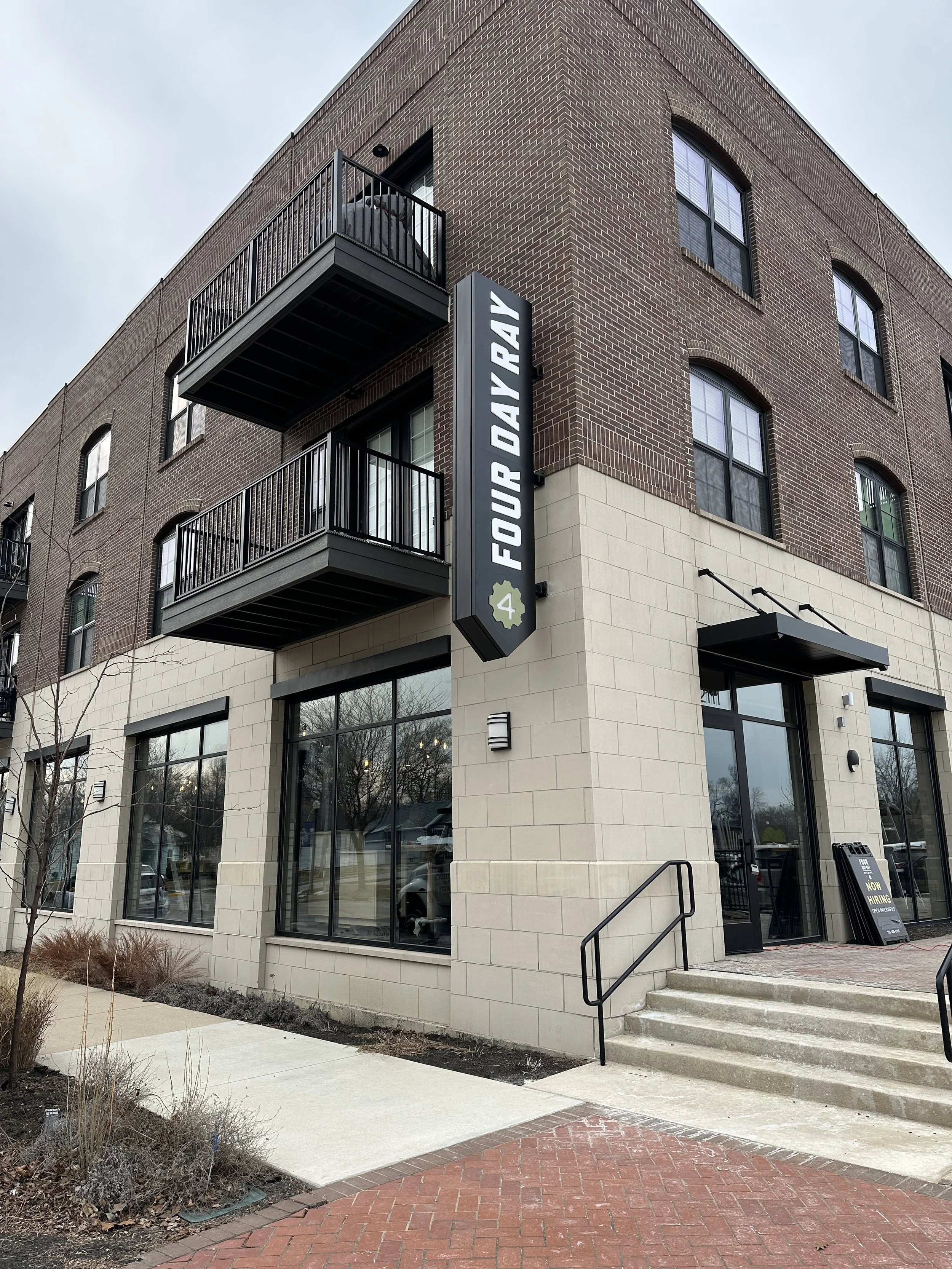 A three-story brick building with black balcony railings and large windows. A vertical sign on the corner reads 'Four Day Ray' with a small gear icon and the number 4. The entrance has steps, glass doors, and a sidewalk with some small plants and tre