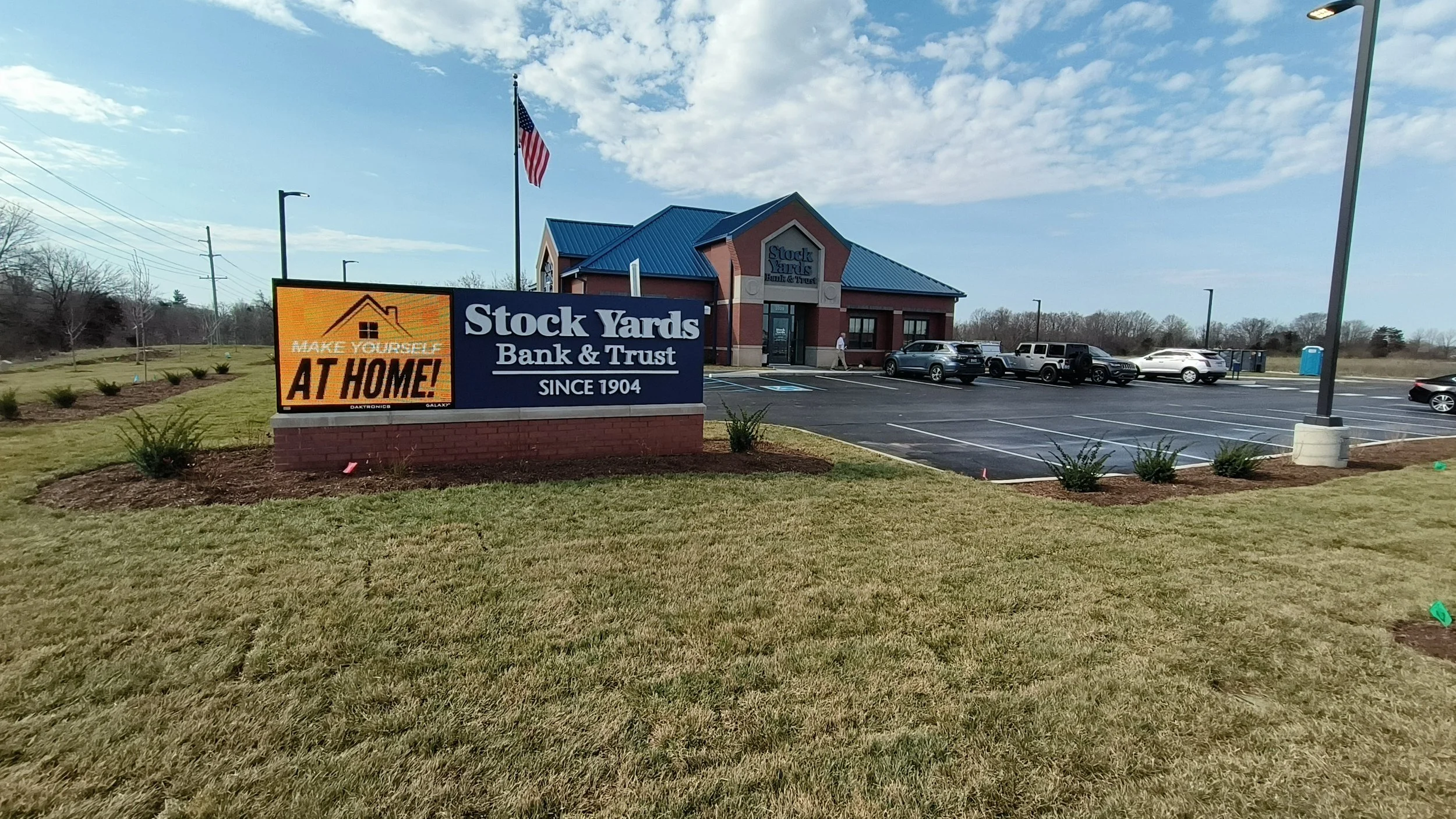 A bank building with a sign that reads 'Stock Yards Bank & Trust since 1904,' and a digital sign that says 'Make yourself at home!' The parking lot has several cars and a portable toilet in the background, with a flagpole flying an American flag. The