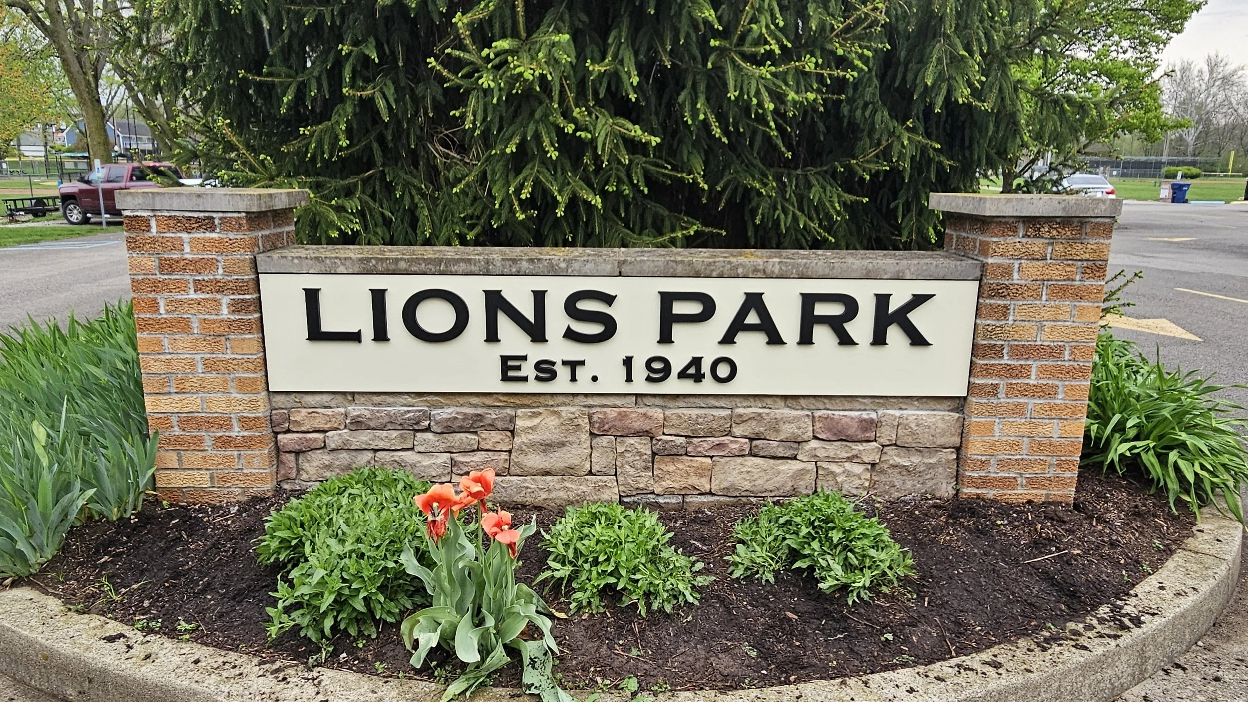Sign for Lions Park, established in 1940, situated in a landscaped area with flowers and shrubs, backed by a large evergreen tree, with parking lot and a pickup truck in the background.