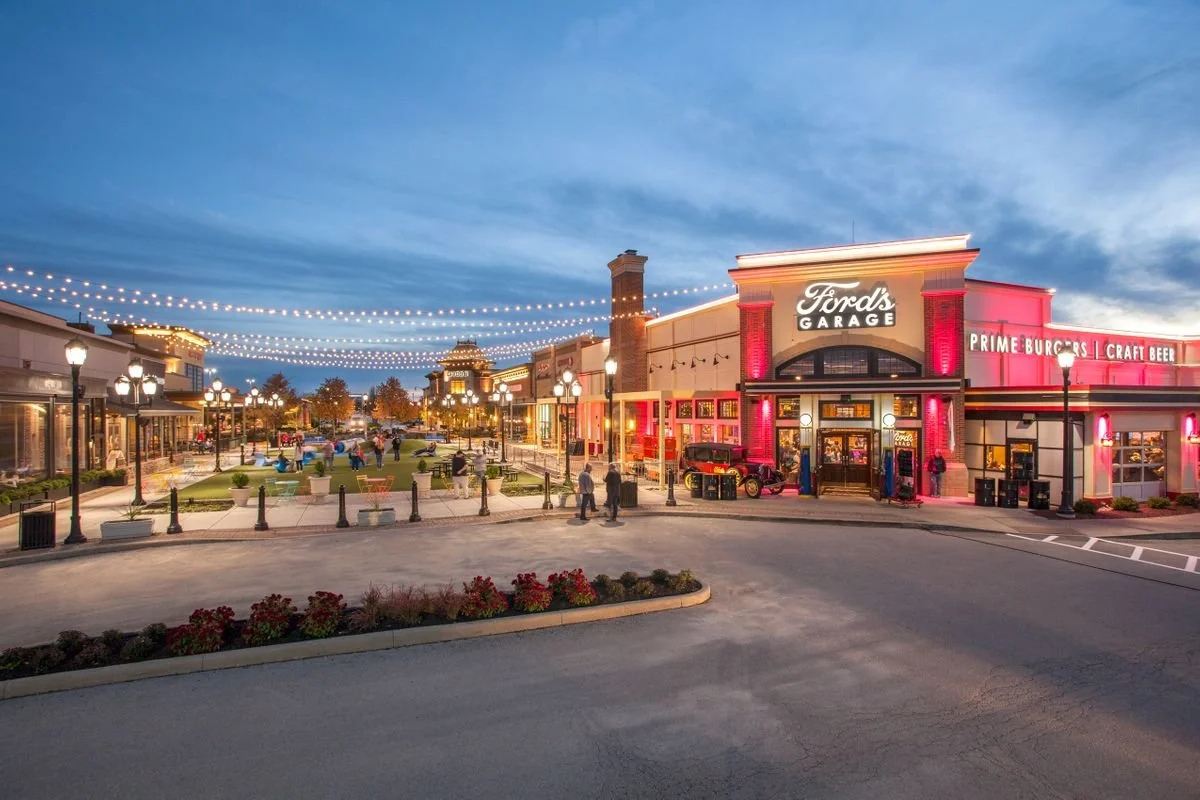 A lively shopping and dining area with a restaurant named Ford's Garage, string lights overhead, and people walking around in the evening.