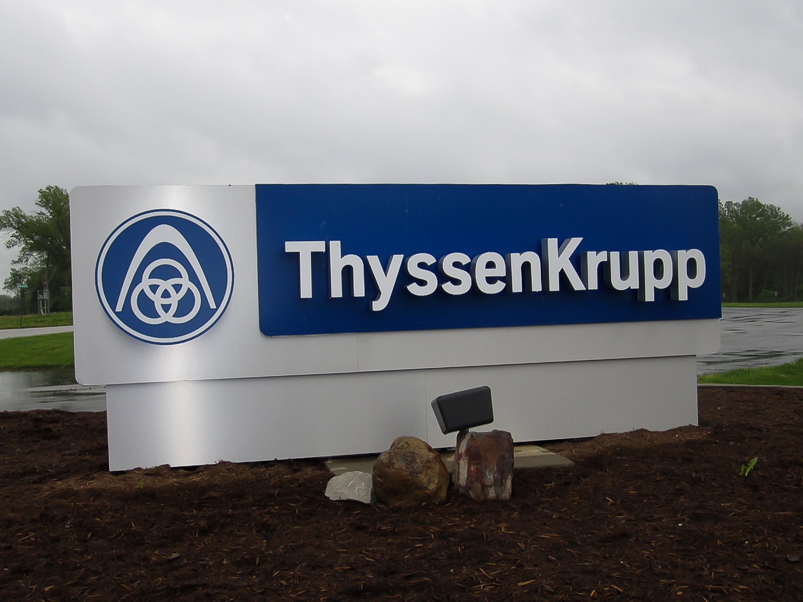 Sign for ThyssenKrupp with company logo, outdoors on cloudy day, surrounded by mulch and small rocks.
