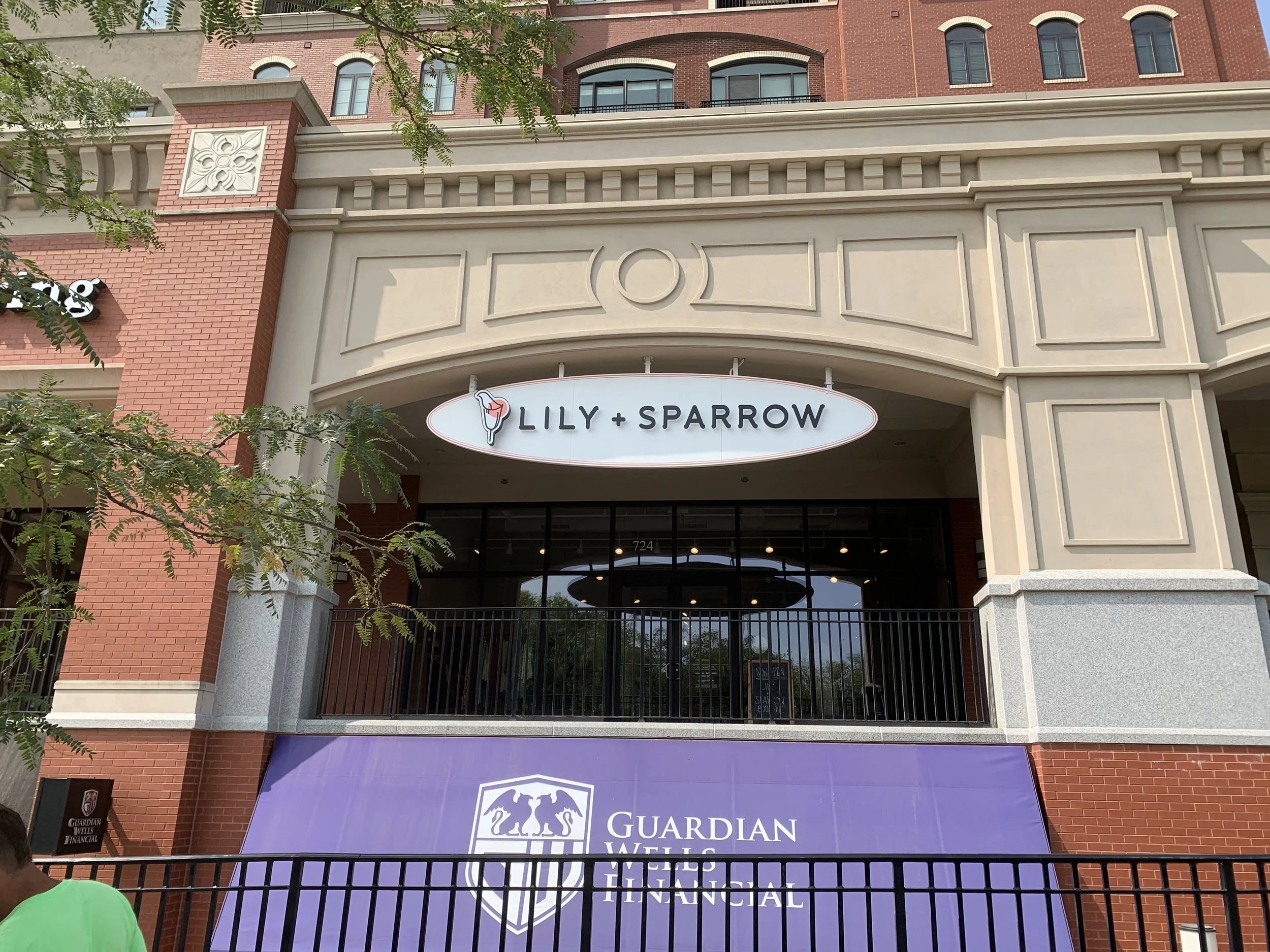 Exterior view of a commercial building with signs for 'Lily + Sparrow' and 'Guardian Wells Financial', featuring a red brick and beige facade, trees, and black metal fencing.