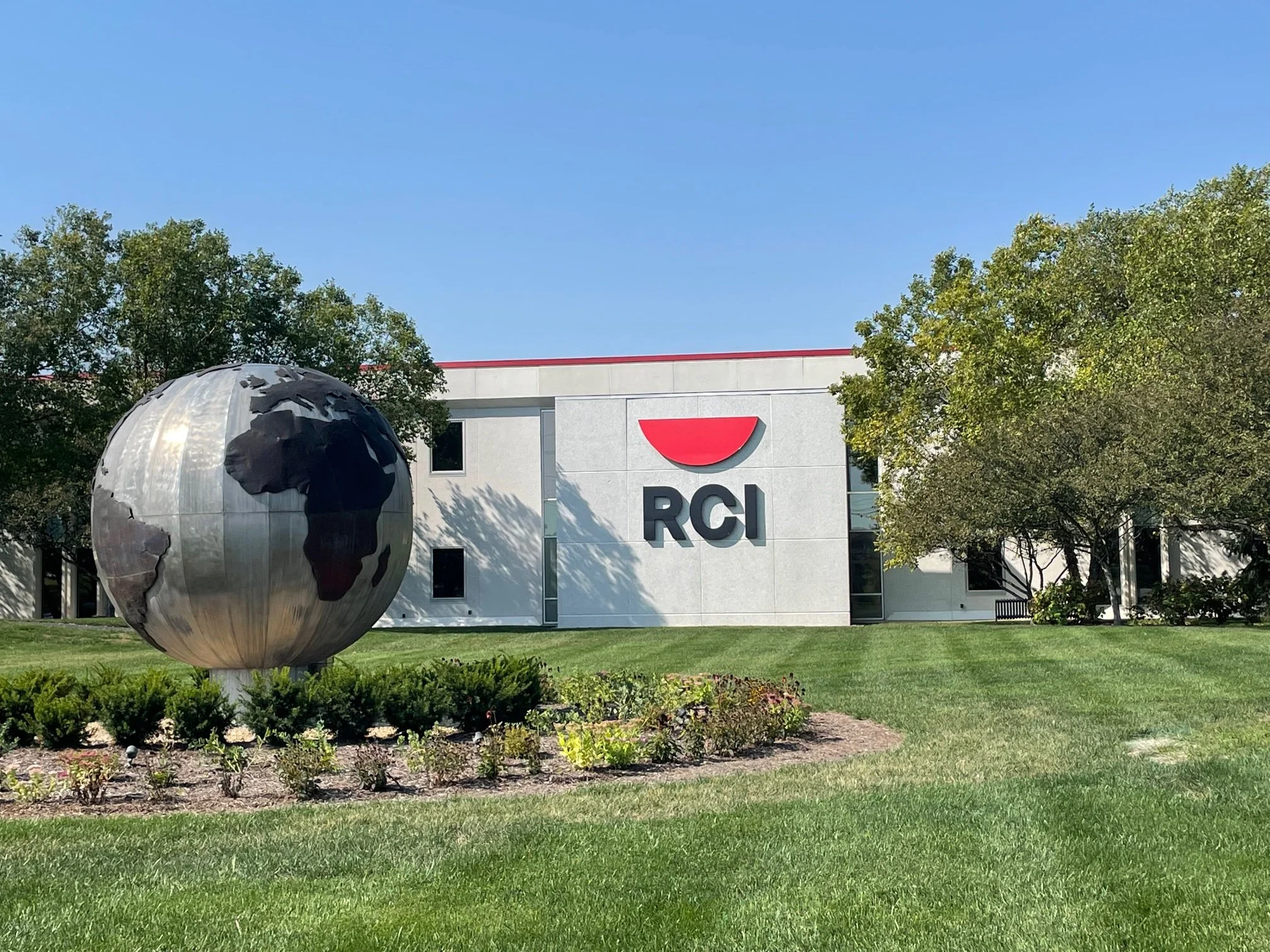 Exterior of a modern office building with a large silver globe sculpture in front, and the RCI logo on the building. There are trees and a well-maintained lawn surrounding the area.