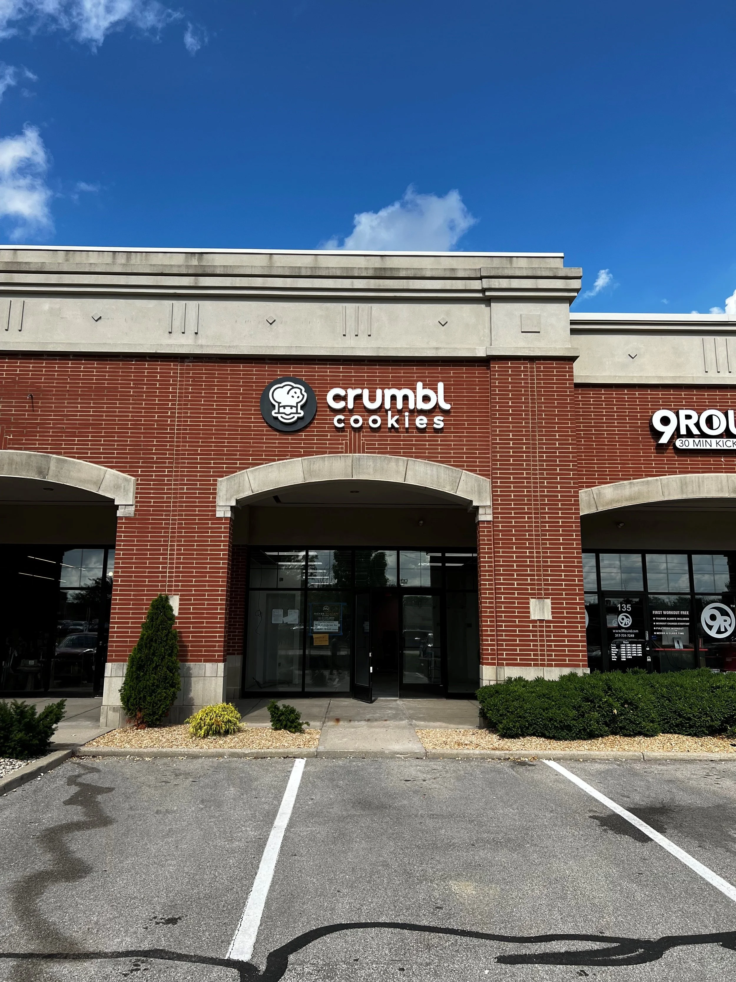 Front of Crumbl Cookies store with parking spaces in the foreground, a brick building, and a blue sky with some clouds.
